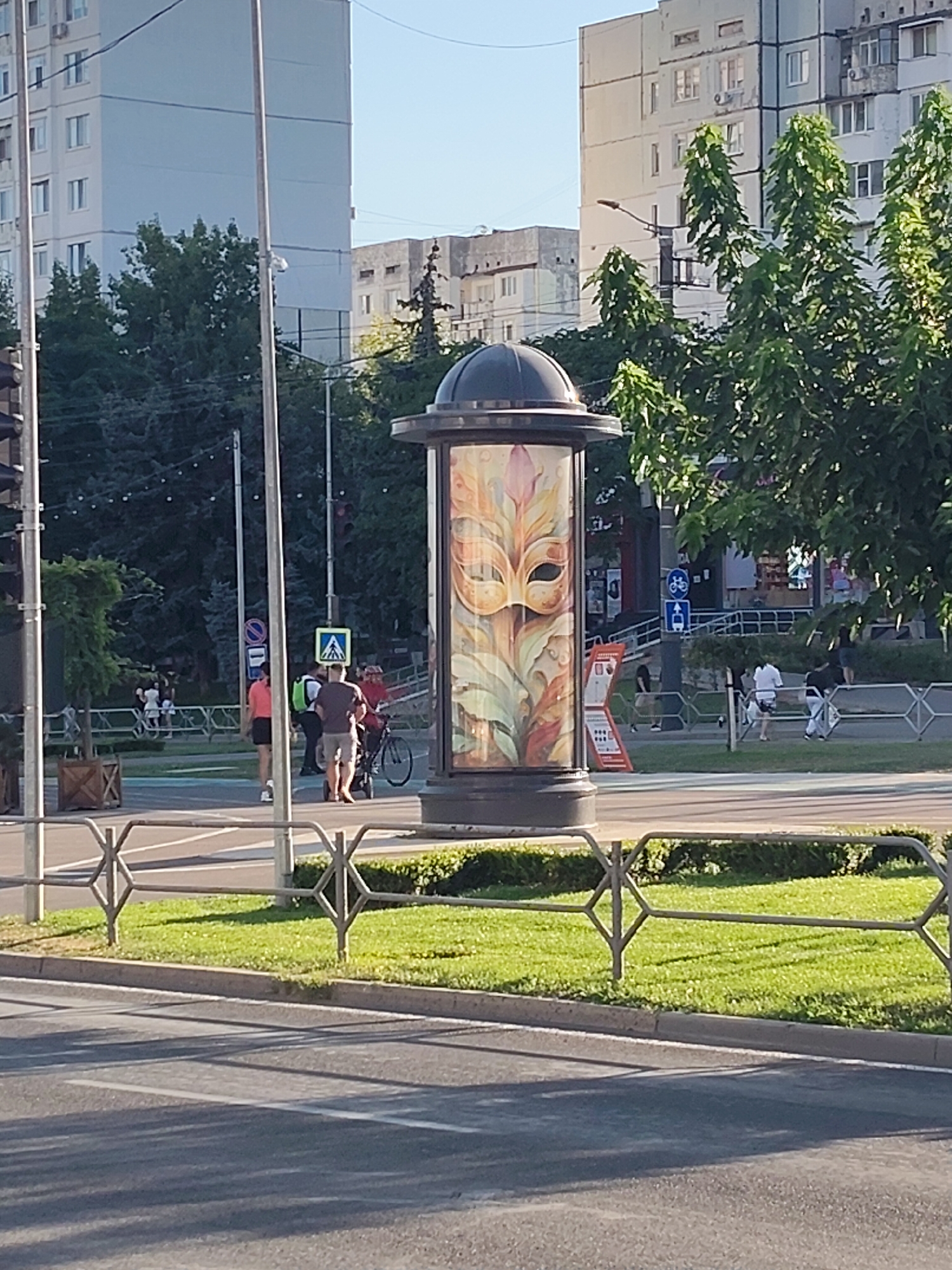 The image depicts a street scene in Chișinău, Moldova, with a prominent cylindrical advertisement pillar displaying a masquerade mask design. The pillar is situated on a sidewalk adjacent to a grassy area and a road. In the background, there are multi-story buildings, trees, and streetlights. Several people are visible in the scene, including individuals walking and a person with a bicycle and a stroller crossing a pedestrian area. The overall impression is a bright, sunny day in an urban environment.