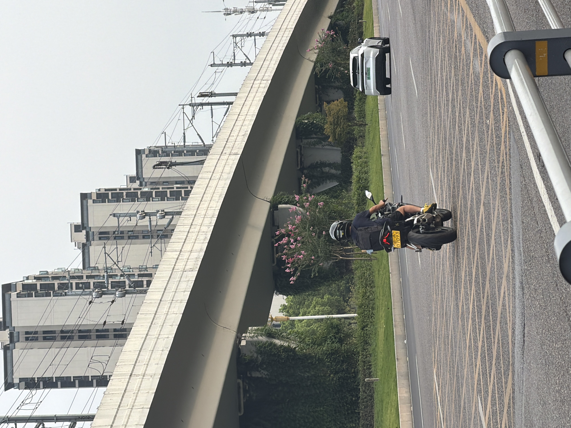A person rides a motorcycle on a city street in Jiangjin, China. The motorcycle is traveling away from the viewer, with a black backpack visible on the rider's back. A white car is driving in the same direction, in a lane to the right of the motorcycle. The street is lined with green grass and trees, with a large elevated structure, likely a bridge or overpass, overhead. The overpass has train tracks and overhead electrical wires. In the background, several modern apartment buildings are visible. The sky is overcast. The street has yellow cross-hatching painted on it, indicating a special lane or intersection.
