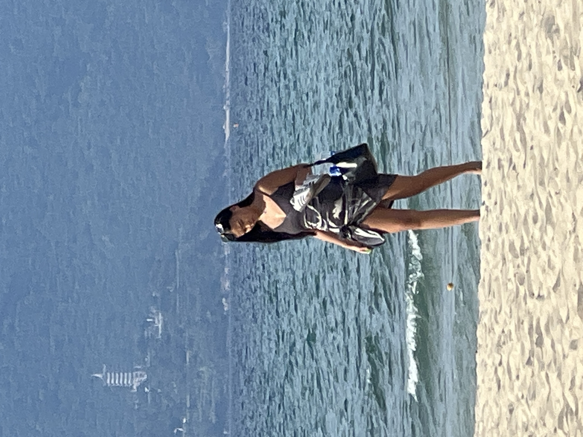 A woman stands on a sandy beach with the ocean in the background. She is wearing a black swimsuit with a patterned wrap skirt, sunglasses on her head, and carrying a tote bag. In the distance, across the water, a mountainous landscape is visible, with a pagoda-like structure on one of the hills. The waves are gentle, and the sky is clear. This scene is consistent with coastal areas in Đà Nẵng, Vietnam, such as My Khe Beach, with views of the Son Tra Peninsula in the distance.