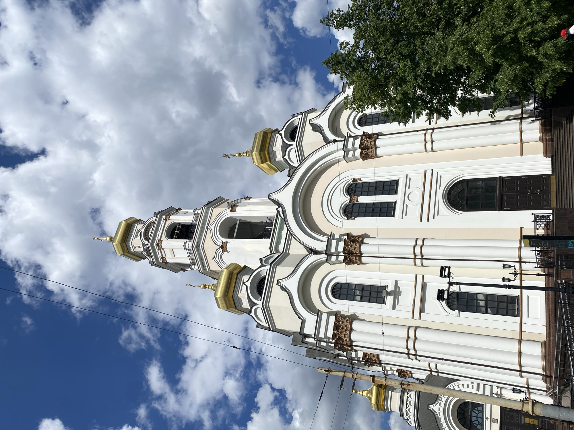 The image shows the Church on Blood in Yekaterinburg, Russia. The view is from the ground, looking up at the church's white walls, gold domes, and some of the surrounding trees. The sky is partially cloudy. There are no people visible. Utility lines are present in the lower left of the frame.