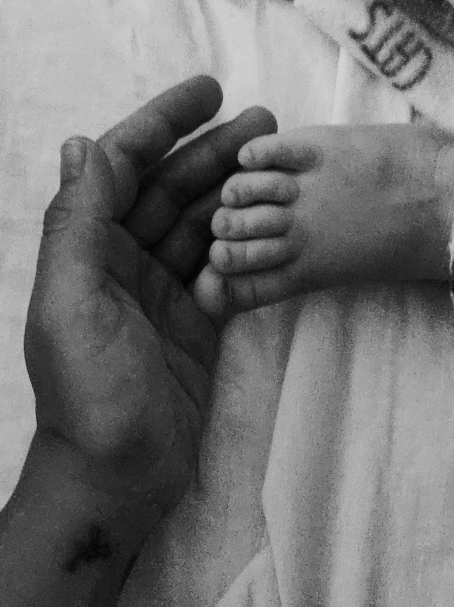 A black and white image shows an adult's hand gently holding a baby's hand. The background appears to be a light-colored fabric, possibly a blanket or sheet. Part of what might be a logo or text, ending in "TED," is visible in the upper right corner. There is no indication of the city of Chaikovsky, Russia.