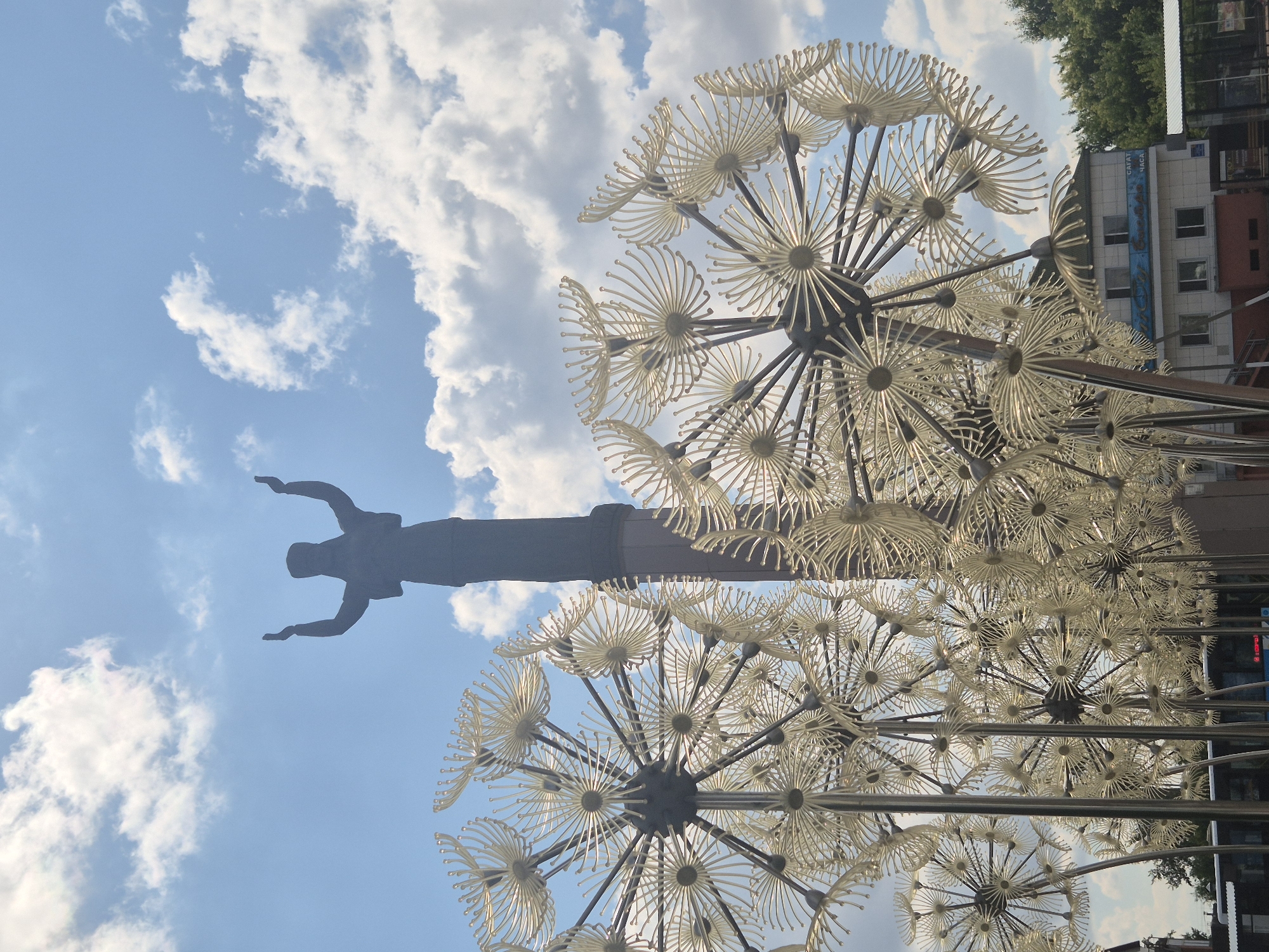 A tall, dark statue of a figure with outstretched arms is silhouetted against a partly cloudy blue sky. In the foreground, a cluster of large, stylized dandelion sculptures made of metal and translucent material are arranged in a dense formation. The sculptures have a central dark sphere with radiating spokes, each ending in a small, petal-like element. Behind the sculptures and the statue, a low-rise building with several windows and a white, arched roofline is visible, along with green trees. The scene appears to be in an outdoor public space in Kokshetau, Kazakhstan, as indicated by the distinctive dandelion sculptures which are a known landmark in the city.