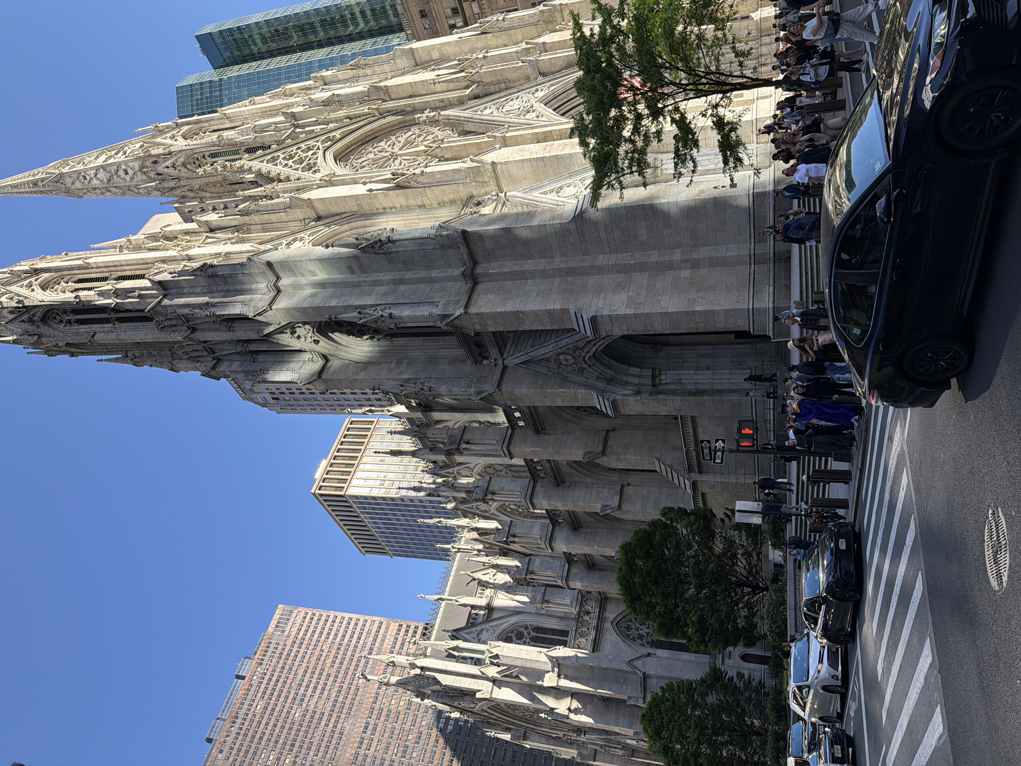 The image depicts St. Patrick's Cathedral in New York City, USA, taken from street level. The gothic-style architecture is prominent, with its spires and intricate stonework reaching towards the blue sky. Several modern high-rise buildings surround the cathedral, juxtaposing the old and new.
On the right side of the image, there's a line of people standing along the cathedral's exterior, likely tourists or visitors. Several dark-colored cars are parked along the curb, near a crosswalk with white diagonal stripes. A traffic light is visible with a red signal illuminated. The street surface is paved and appears to be asphalt. Trees are interspersed along the street.