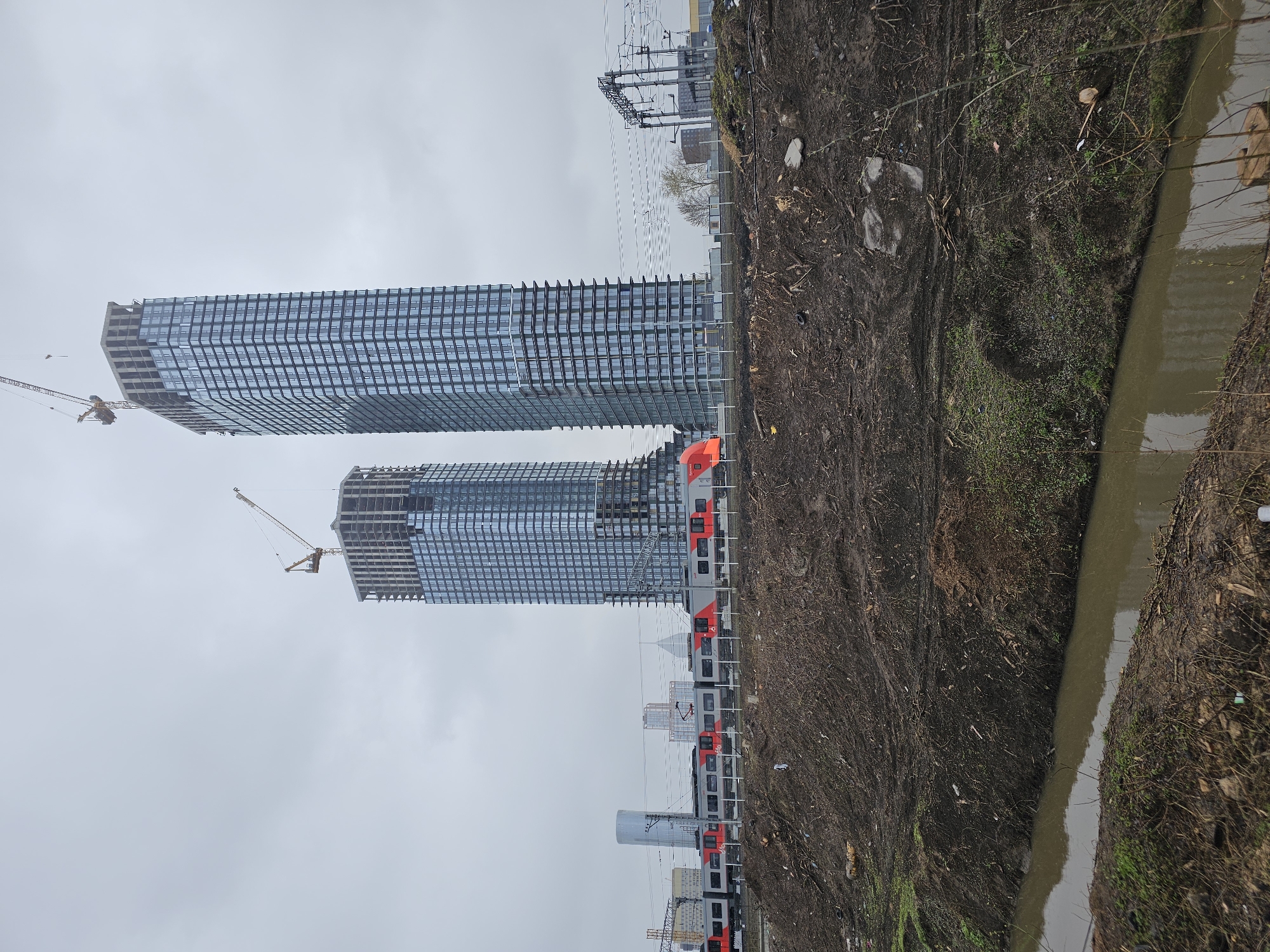The image shows a construction site in Khimki, Russia, with two tall, modern buildings under construction. Cranes are visible near the top of the buildings, indicating ongoing construction. A train is passing by along the construction site. There is a muddy area in the foreground, with a narrow, water-filled trench or drainage ditch running alongside it. The sky is overcast, suggesting a gray day.