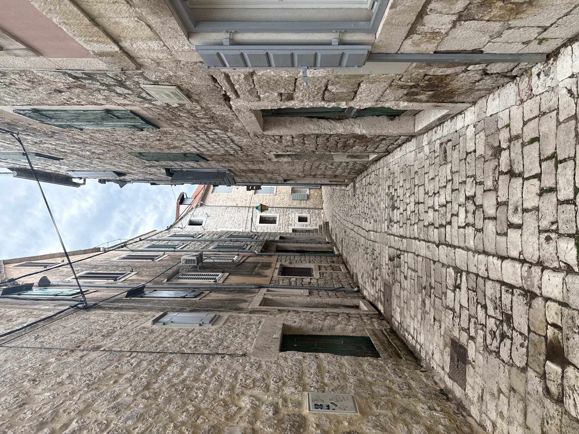 This is a ground-level, eye-level, medium shot of a narrow cobblestone alleyway in the historic town of Stari Grad, Croatia. The alley is flanked by tall, ancient stone buildings. The stone construction is visible on both sides of the alley, with rough-hewn blocks forming the walls. Many windows are present, some with weathered, louvered shutters, predominantly in shades of green and grey. The cobblestone pavement is uneven and shows signs of age and wear, with some sections appearing damp or wet. There are various architectural details, including doorways, drainpipes, and electrical conduits running along the building facades. A street lamp is visible in the distance, illuminating a portion of the alley. The sky above is overcast and cloudy. No people are present in the scene. The overall impression is one of a well-preserved, historic urban environment.