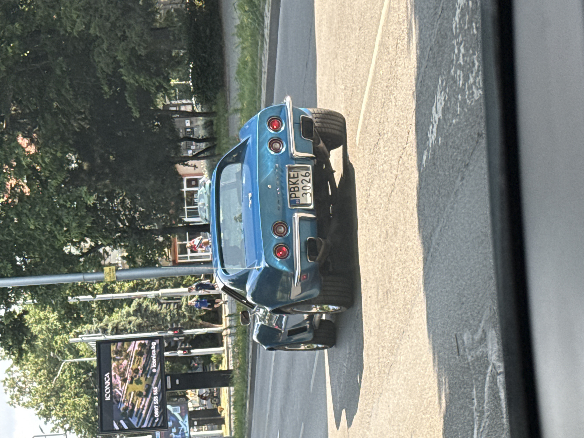 The image depicts a blue Chevrolet Corvette C3, model year approximately 1968-1972, traveling away from the viewer on a paved road in Plovdiv, Bulgaria. The vehicle bears a Bulgarian license plate "PBKE 3026," with "PB" signifying Plovdiv. The car features dual circular taillights, chrome bumper, and wide rear tires.
The scene is an urban environment under clear daylight, casting distinct shadows. To the left of the road, a wide sidewalk is visible, bordered by trees and street infrastructure. A large black billboard displays the brand "ICONICA" and a rendering of a planned development, along with the phone number "0897 555 555" and website "iconica.bg." Below it, a smaller billboard is partially visible. Several pedestrians are on the sidewalk: two individuals, one in a blue shirt raising an arm, walk away from the viewer near the billboard. Further back, a person is near a bicycle.
To the right of the road, trees and foliage line the thoroughfare, partially obscuring background buildings. A metal utility pole with an illegible yellow sign is present. Two additional individuals are partially visible on the far right, one in a red shirt and another in a dark shirt. The road appears to have multiple lanes, and the overall setting is characteristic of a city street or boulevard.