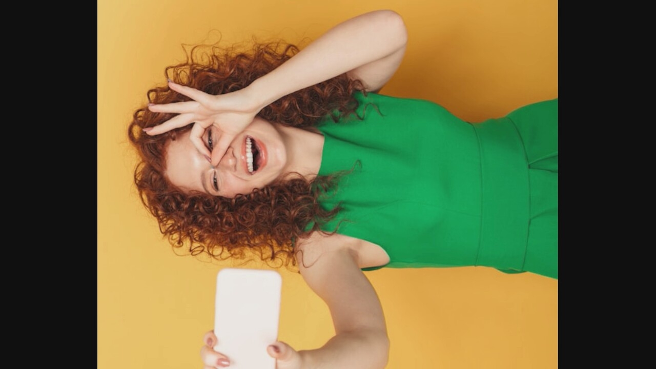 A young woman with curly red hair, wearing a bright green dress, is holding a white smartphone up to take a selfie. Her left eye is covered by her fingers, and she is smiling broadly. The background is a solid yellow color.