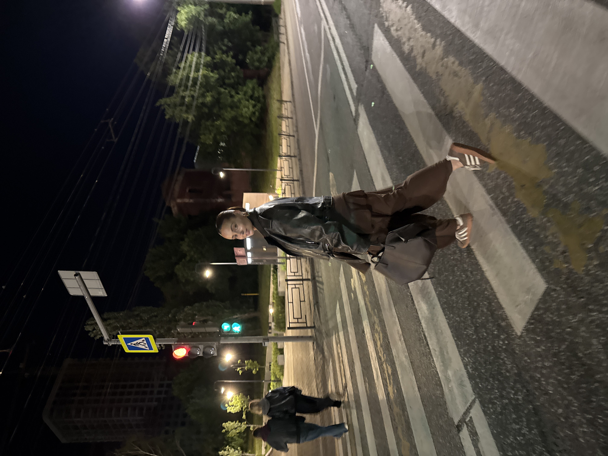 The image shows a woman standing on a pedestrian crossing at night. She is wearing a dark jacket and brown pants, and has a dark bag over her shoulder. The traffic light is red. Behind her are two other people walking on the crosswalk. The location appears to be Izhevsk, Russia.
