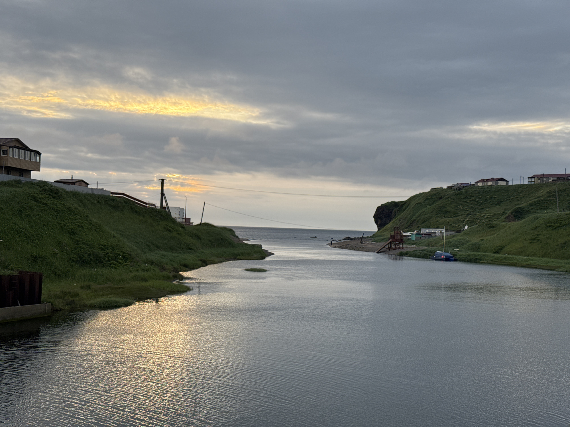 The image depicts a serene coastal scene in Kuril'sk, Russia, under a cloudy sky with a band of golden light breaking through near the horizon. A river or inlet flows into the ocean, flanked by green, grassy hills dotted with a few small houses. On the left hill, a multi-story building with a sunroom extension is visible. On the right hill, several houses and a small dock with a blue boat are present. The water's surface is calm with ripples reflecting the subdued light. A solitary figure can be seen walking near the shore on the right side of the inlet. The overall atmosphere is tranquil and suggests a remote, natural environment.