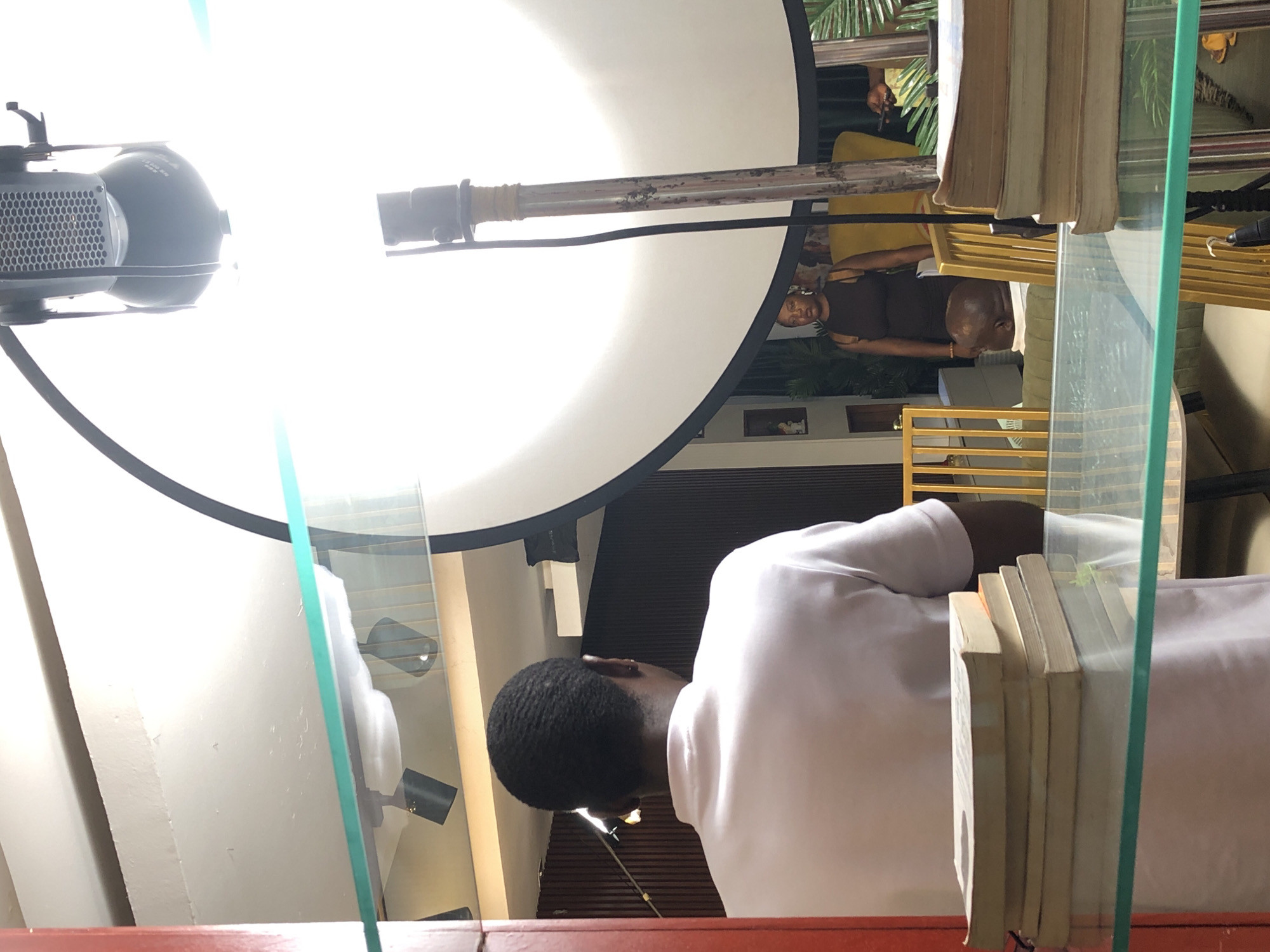 The image depicts an indoor scene, likely a professional photoshoot or video recording setup. The perspective is taken from slightly above and behind a person, looking towards a brightly lit area. The image appears to be rotated 90 degrees clockwise, showing the scene vertically.
In the foreground, towards the bottom right, the back of a person's head with short, dark hair, wearing a white shirt, is visible. This person appears to be looking downwards. A transparent glass partition or shelf diagonally crosses the lower section of the frame, and a stack of light-colored books rests on it in the bottom right corner.
The dominant feature of the mid-ground, occupying the entire left side of the image, is a large, circular white light diffuser or reflector with a black rim. A professional studio light with a textured grille is attached to a stand on the far left, emitting a strong white light that illuminates the diffuser. Text reading "HEDLER" and "1000 W" is visible on this light fixture. A metal pole extends horizontally across the center of the diffuser.
In the background, behind the diffuser and glass partition, several elements are discernible. A person wearing a dark top is standing, facing towards the left side of the frame. Their reflection is faintly visible on the surface of the large white diffuser. Further back, parts of another person wearing a yellow garment, green tropical foliage, yellow railing, and interior architectural elements like white walls and dark horizontal slatted blinds or shelves are visible.
The scene is brightly lit by artificial light, suggesting a controlled environment. The presence of professional lighting equipment indicates that an event like a media production is underway. Reflections on the glass and the diffuser add complexity to the composition. The time of day is not explicitly clear but appears to be well-lit, possibly daytime, enhanced by artificial lighting.