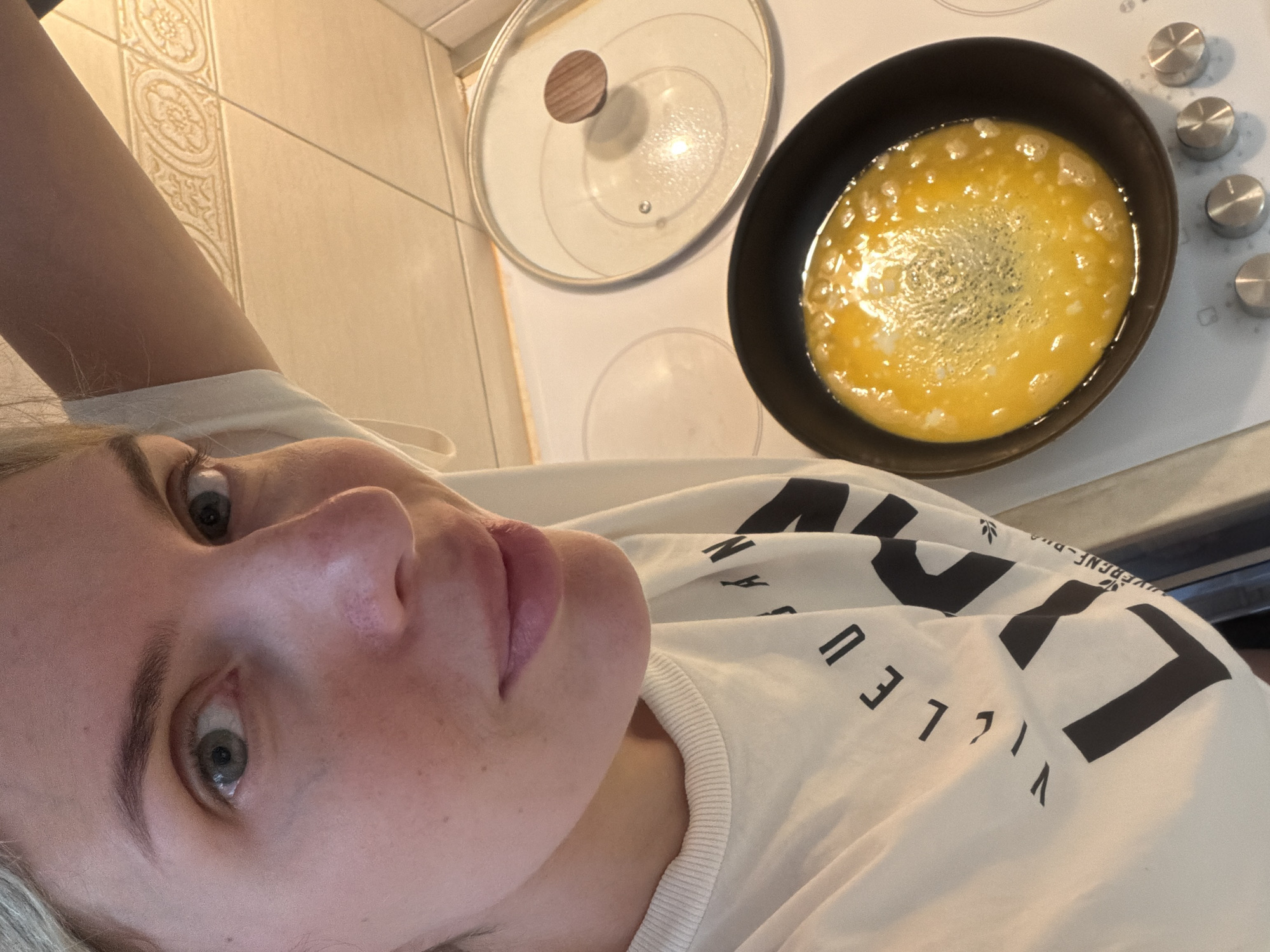The image shows a close-up, low-angle selfie of a young woman with blonde hair. She is wearing a white t-shirt with black text that appears to be a logo or brand name. The woman has light-colored eyes and fair skin with some freckles. Her lips are slightly parted, and she has a neutral expression.
In the background, a black frying pan filled with cooking eggs is visible on a stovetop. Next to the pan, a glass lid with a wooden handle rests on the stovetop. The stovetop has control knobs. The wall behind the stovetop is tiled with a decorative pattern. The lighting suggests it is daytime. The overall setting appears to be a kitchen. The text on the shirt is partially visible, showing letters like "V", "I", "L", "L", "E", "U", and "E", as well as other symbols. The context provided indicates the location is Budva, Montenegro.