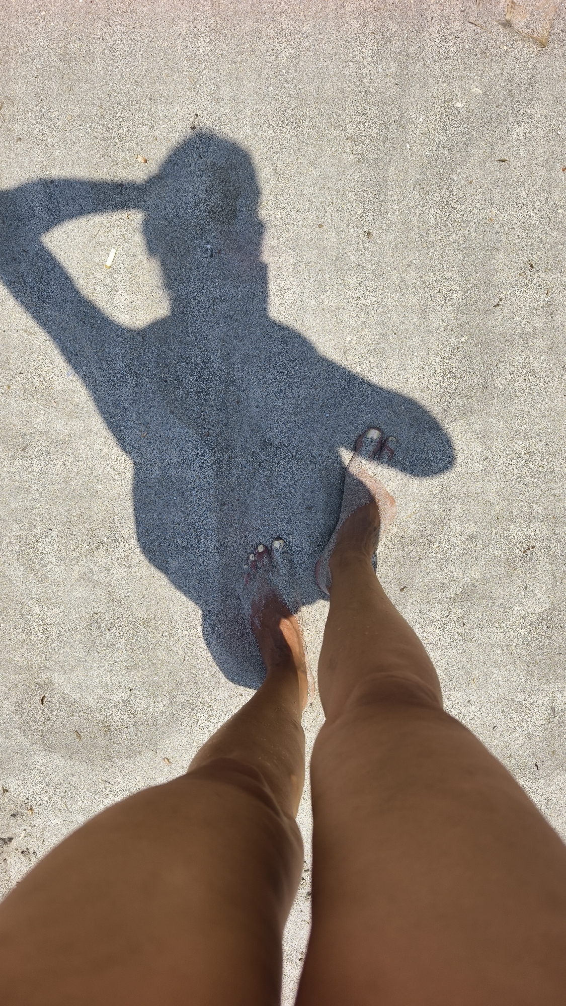The image displays a top-down view of a person's legs and feet standing on a sandy surface, likely a beach. The person's shadow is cast on the sand, showing a figure with an arm raised behind their head. The sand appears light-colored and fine-grained, with a few small, dark specks scattered across it. The person's legs are tanned, and their feet are bare, with toenails painted a light color. The lighting suggests it is daytime, with a strong light source casting a distinct shadow. There are no visible buildings, landmarks, or indications of the specific location within Kuşadası, Turkey. The scene is devoid of any other individuals or objects apart from the person and the natural sand environment.