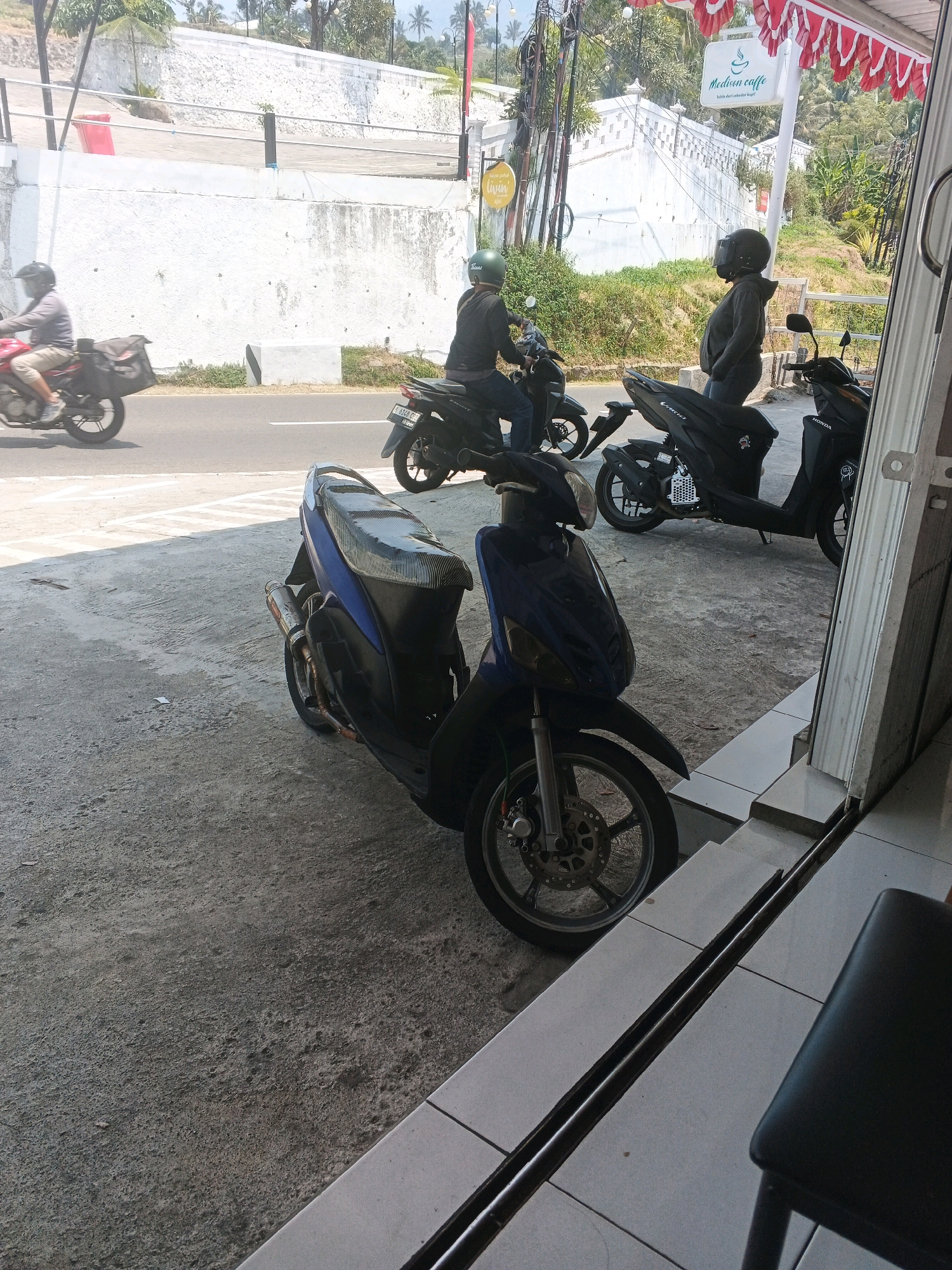 A street scene in Pakopen, Indonesia shows several motorcycles parked and in motion. In the foreground, a dark blue scooter with a carbon fiber patterned seat is parked on a concrete surface. Behind it, two black scooters are also parked. One has a license plate that reads "Y 6743 CZ". Further back, on the asphalt road, a person on a red motorcycle with a black bag attached to the back is moving from left to right. Another person on a black motorcycle with a green helmet is also seen on the road. In the background, a white wall and lush greenery are visible. A sign for "Medison caffe" hangs from a pole. The scene appears to be a public area with a café or shop entrance to the right.
