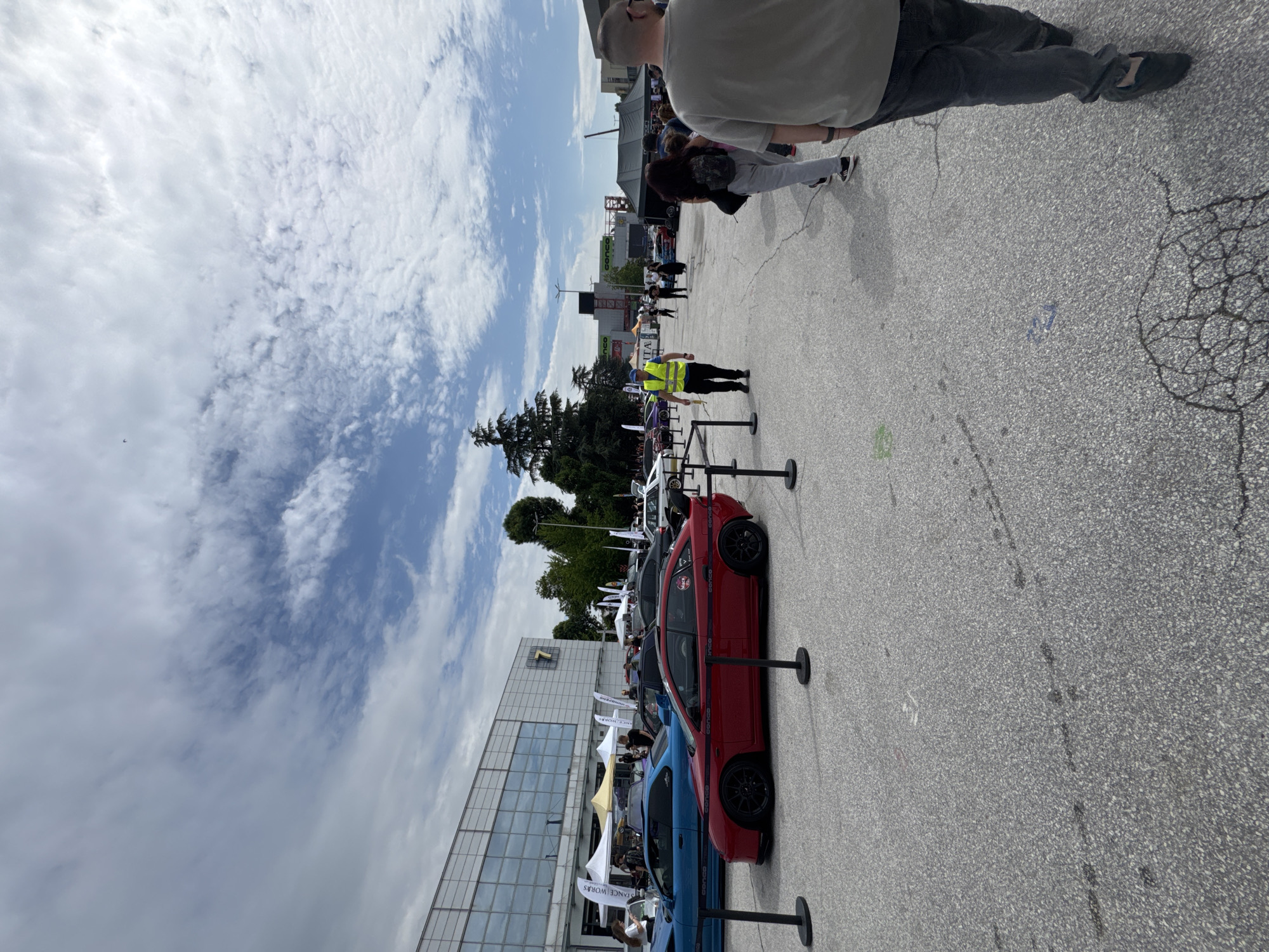 The image depicts a car exhibition in Plovdiv, Bulgaria. Several modified cars are parked in a row, separated by metal posts. A person wearing a yellow vest stands nearby. A building with glass windows is to the left of the cars. The sky is cloudy. People are present, some near the cars and others in the background. The ground is paved.