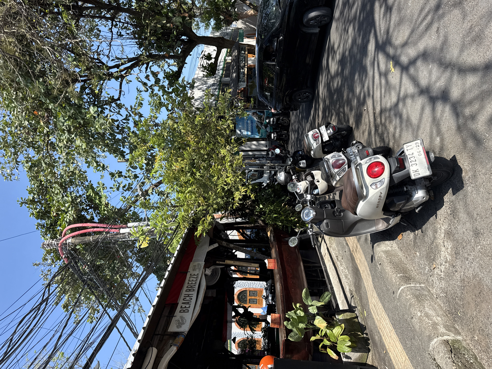 The image depicts a street scene in Denpasar, Indonesia, on a sunny day. In the foreground, several scooters are parked along the sidewalk. The closest scooter is a white Honda Scoopy with a brown seat and a red taillight. Its license plate reads "DK 3234 TY" and "06-29". Behind it, another scooter, also white and brown, is partially visible. Further down the street, more scooters are parked.
To the left, a building with an open-air structure resembling a restaurant or cafe is visible. A sign above the entrance reads "BEACH BREEZE". The establishment has wooden counters, potted plants, and what appear to be tables and chairs inside.
A black car is parked on the right side of the street, partially obscuring a building behind it. Trees with lush green foliage line the street, casting dappled shadows on the pavement. Overhead, a complex network of electrical wires is strung between poles, with some red conduits visible. The sky is clear and blue. No people are clearly visible in the image.