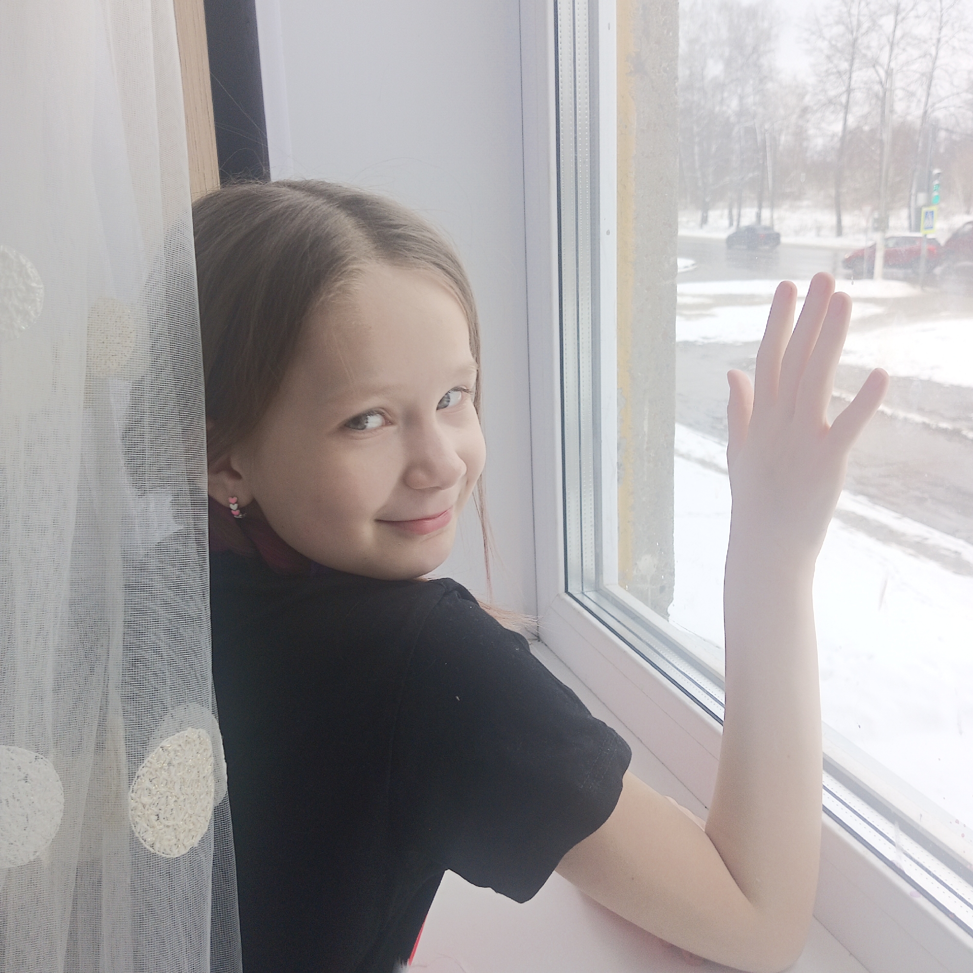 A young girl with light brown hair stands by a window, her left arm raised with her hand outstretched towards the glass, possibly waving. She wears a black shirt and heart-shaped earrings. The window overlooks a snow-covered street with cars and a pedestrian crossing sign visible. A white curtain with dotted patterns hangs to the left of the girl. The scene appears to be in Chaikovsky, Russia, based on visual markers such as architectural style and signage.