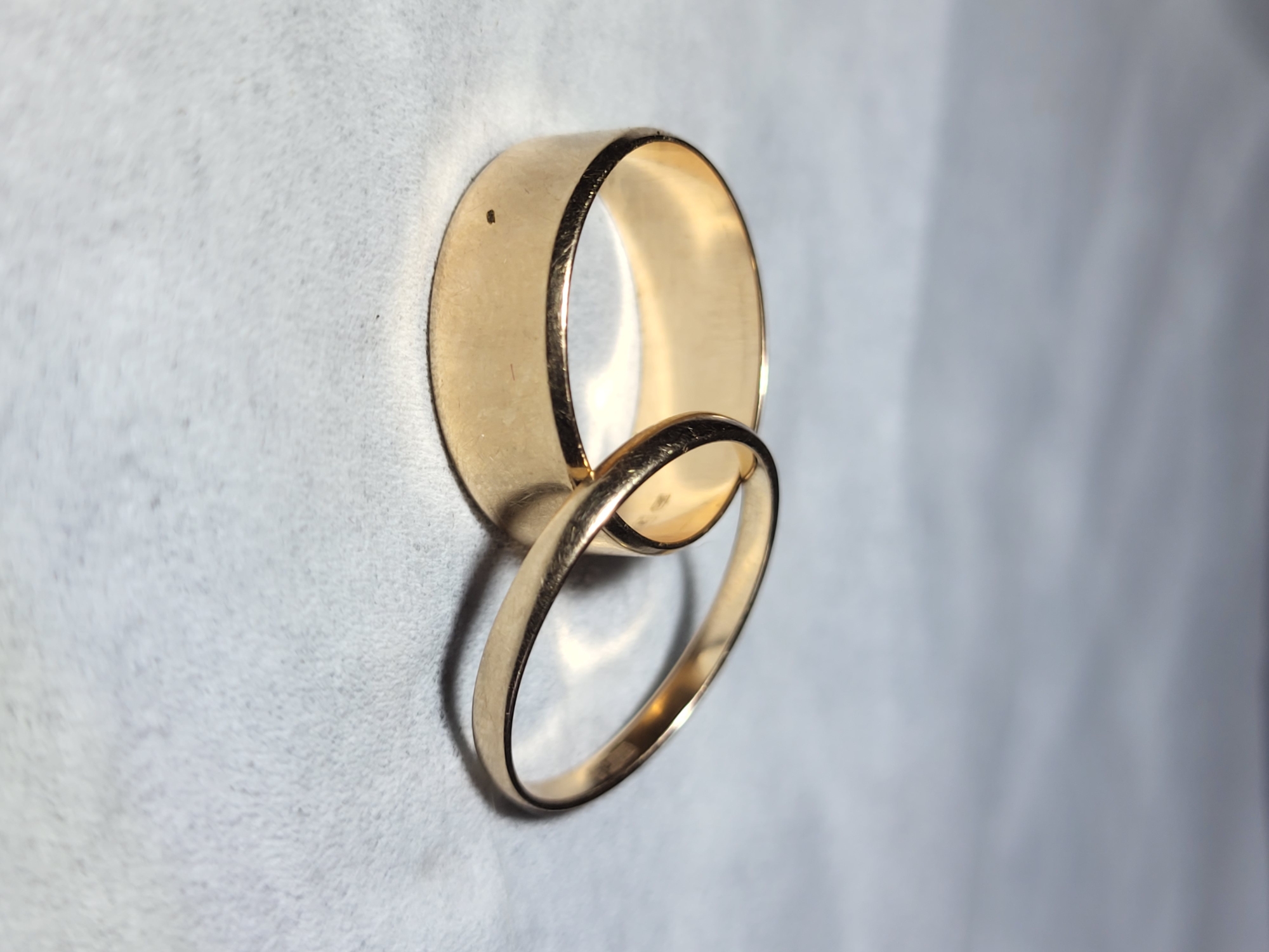 Two gold rings are resting on a light-colored, textured surface. One ring is wider and positioned closer to the foreground, with its band resting horizontally. The second ring is narrower and lies diagonally across the first ring, slightly overlapping its edge. The rings are made of polished gold and reflect light. The background is out of focus, with soft, muted tones suggesting a diffused light source. There is no discernible indication of the city of Horishni Plavni, Ukraine, or any specific urban environment. The scene is static and devoid of any human presence or action.