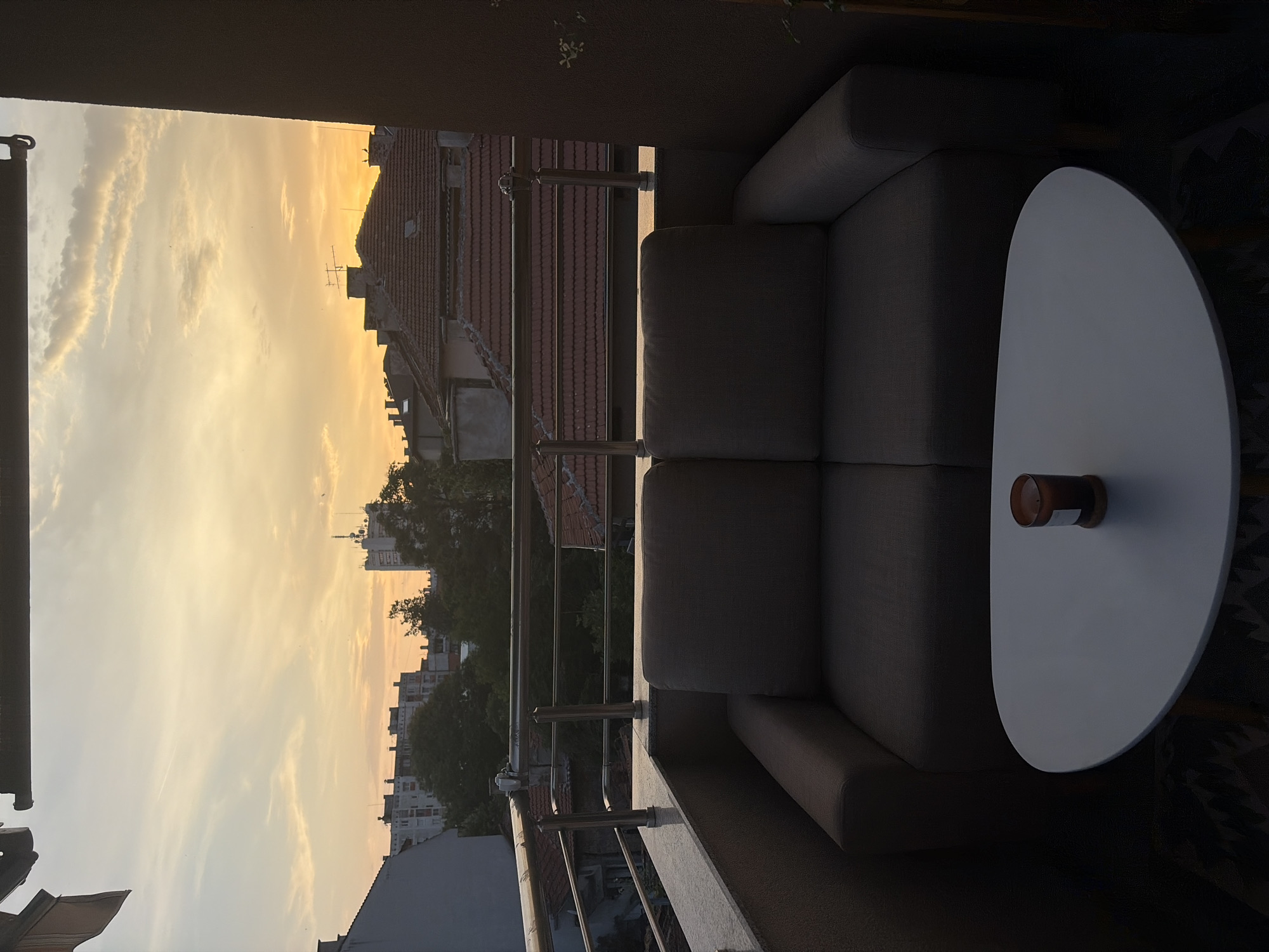 This image depicts a balcony scene at sunset, oriented vertically. The foreground features a modern, gray sectional sofa and a white oval coffee table. On the table, a cylindrical object, possibly a candle or diffuser, is visible. The background reveals a city skyline with buildings and rooftops bathed in the warm, golden light of the setting sun. The sky is filled with soft, wispy clouds. The scene suggests a peaceful, relaxing atmosphere, likely an outdoor seating area. The time of day is clearly sunset, indicated by the colors of the sky and the soft lighting. There are no people visible in the image. No text is discernible.