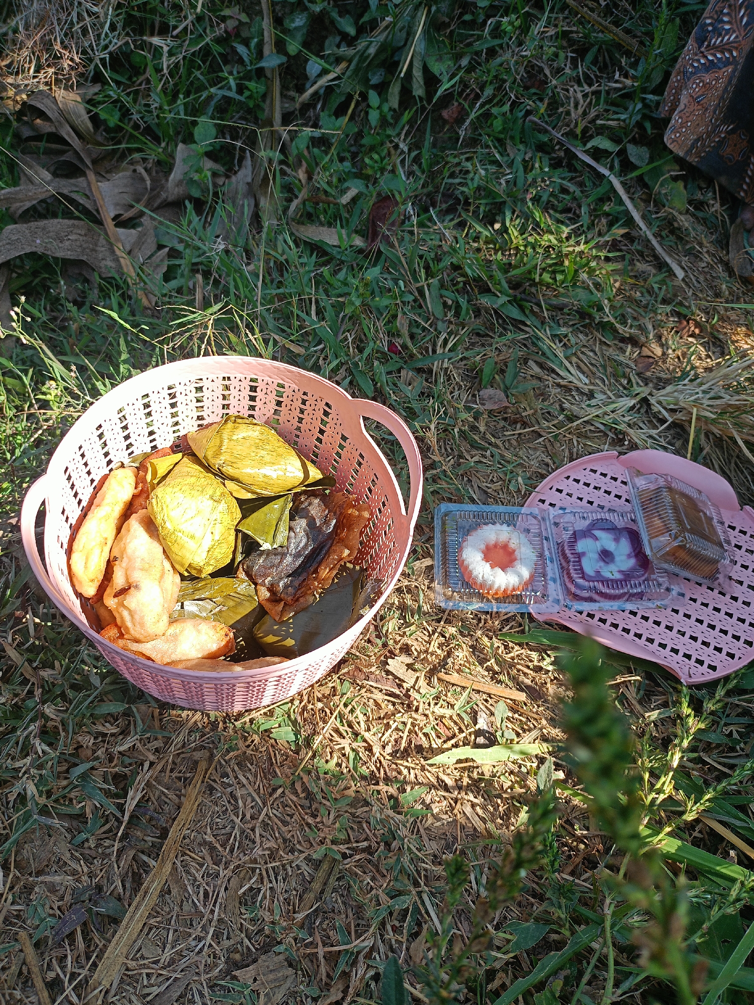 The image depicts a picnic scene set outdoors on grass and dry leaves. In the foreground, a pink woven basket is filled with various food items, including what appear to be fried bananas and some food wrapped in banana leaves. To the right of the basket, on a pink woven mat, are three small plastic containers of sweets. The environment suggests a natural setting, possibly a park or a field, with sunlight casting shadows. The background shows more grass and foliage. The scene is set in the city of Jelbuk, Indonesia.