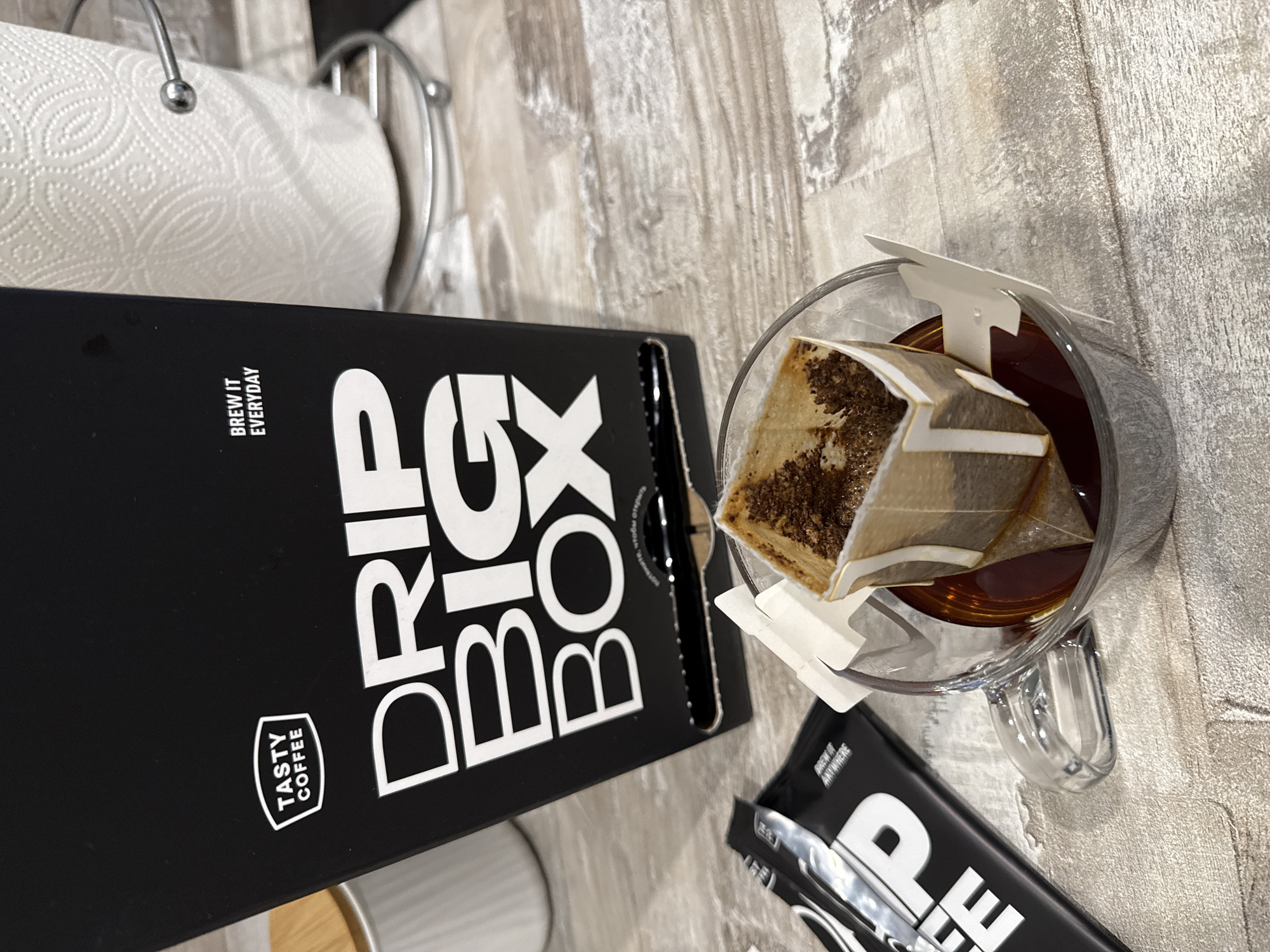 A top-down shot shows a "Drip Box" of "Tasty Coffee" alongside a clear glass mug with a drip coffee bag inside it, sitting on a gray wood-like surface. A roll of paper towels is in the background on the left. A smaller packet of coffee is also on the surface. The text on the box reads, "Brew it Everyday." The cup appears to contain coffee. The packet of coffee reads "Brew It Anywhere." There is no indication of the location of Izhevsk, Russia in the image.