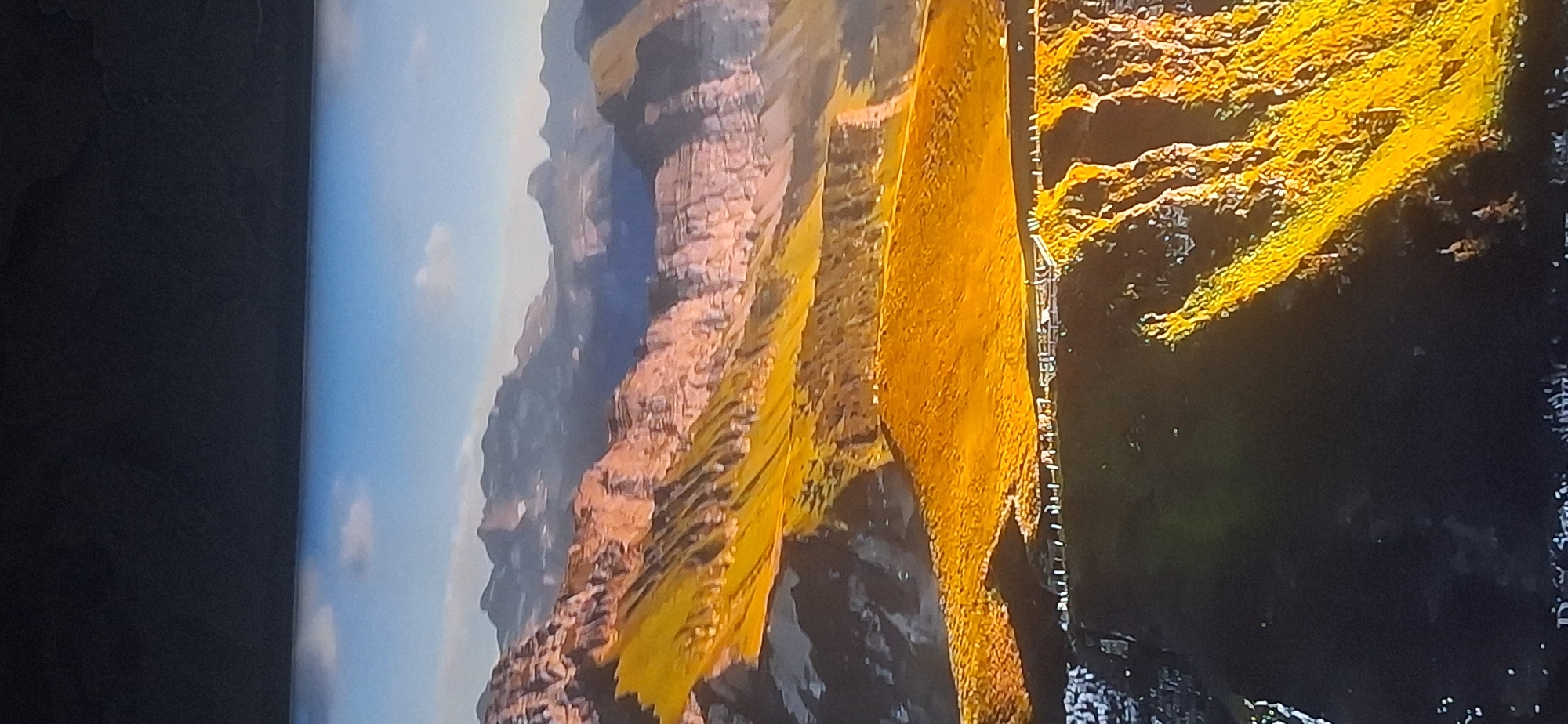 The image shows a panoramic view of a mountainous landscape. The foreground features a steep, dark slope with scattered patches of bright yellow and green vegetation illuminated by sunlight. A white railing or fence runs along the edge of this slope, separating it from a bright, golden-hued field further back. This field slopes upwards towards a series of rugged, multi-layered mountains. The mountains exhibit a range of colors, from warm oranges and yellows on their sunlit faces to cooler blues and purples in their shadows and distant peaks. The sky above is a clear, light blue with a few wispy white clouds. The scene is devoid of any human presence, vehicles, or urban structures. The location of Bila Tserkva, Ukraine is not depicted in this image.