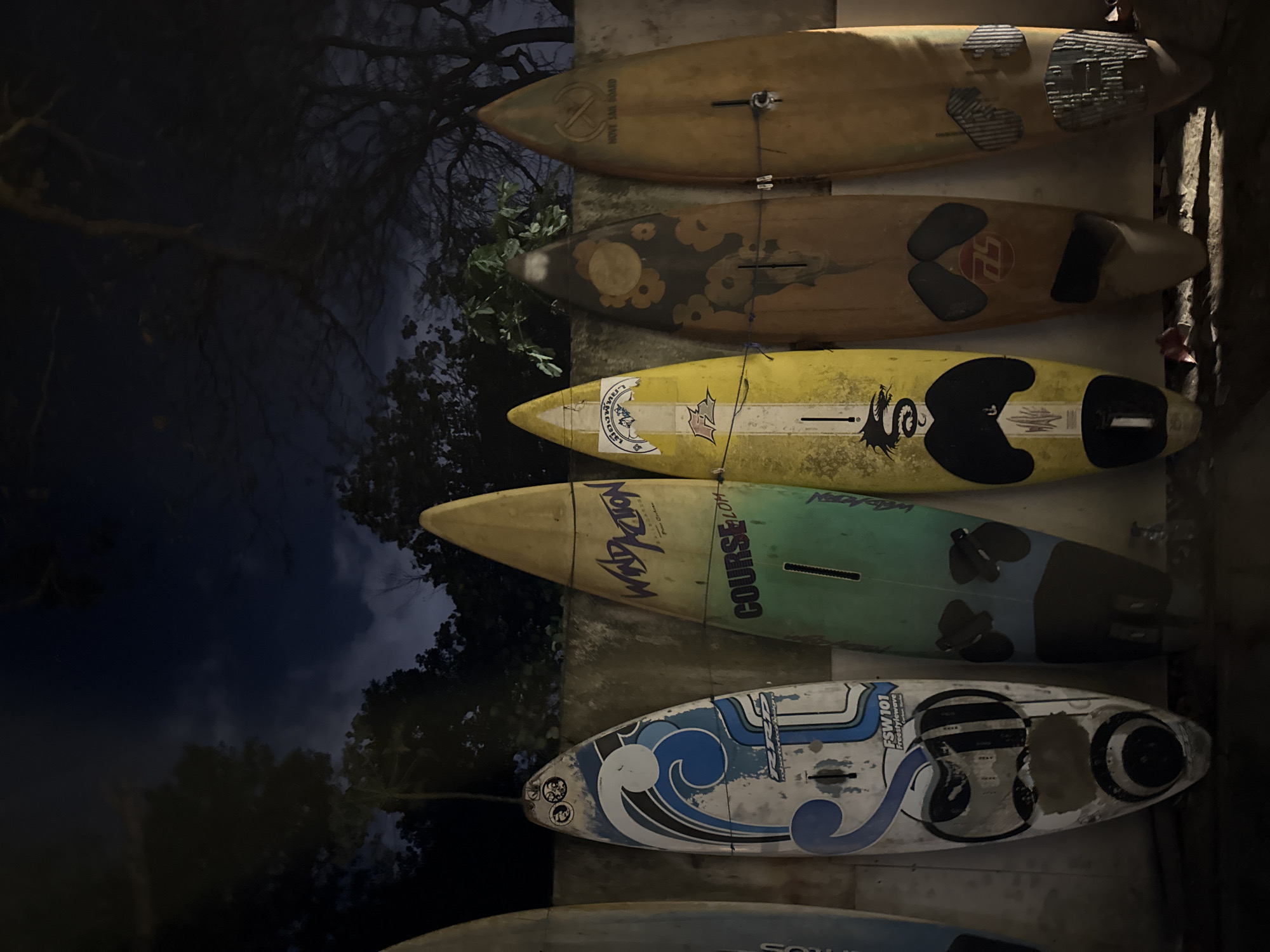 A dimly lit, outdoor scene at night shows a row of five surfboards leaning against a concrete wall. The surfboards are various colors and designs. From left to right, the first surfboard is partially visible and appears to be blue. The second surfboard is white with blue wave graphics and the text "FSW 101 Freestylewave." The third surfboard is green and yellow with text that reads "WindAction" and "COURSE LOH." The fourth surfboard is yellow with a black dragon graphic and a white stripe. The fifth surfboard is brown with a floral pattern. The sixth surfboard is yellow with a circular emblem and text that reads "MOVE ON BOARD." The ground in front of the surfboards is covered in shadows. The background consists of dark trees and a dark sky with some faint clouds. The overall impression is one of a storage area for sporting equipment, possibly a surf shop or rental location, in Denpasar, Indonesia.
