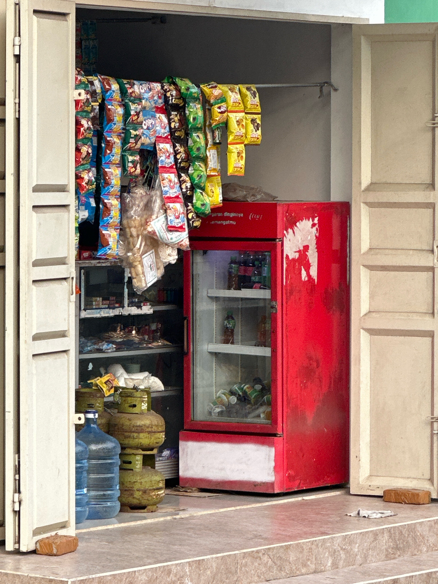 The image shows a small, open-air convenience store, possibly located in Saruni, Indonesia. The storefront has two large, white double doors that are open, revealing the interior. Inside, a red refrigerator is visible, filled with drinks. Shelves on the left are stocked with various goods, including snacks and bottled water. There are also propane tanks and a brick in front of the store. The scene appears to be outdoors, with the store set up on a paved area with stairs leading up to it. The lighting suggests it is daytime.