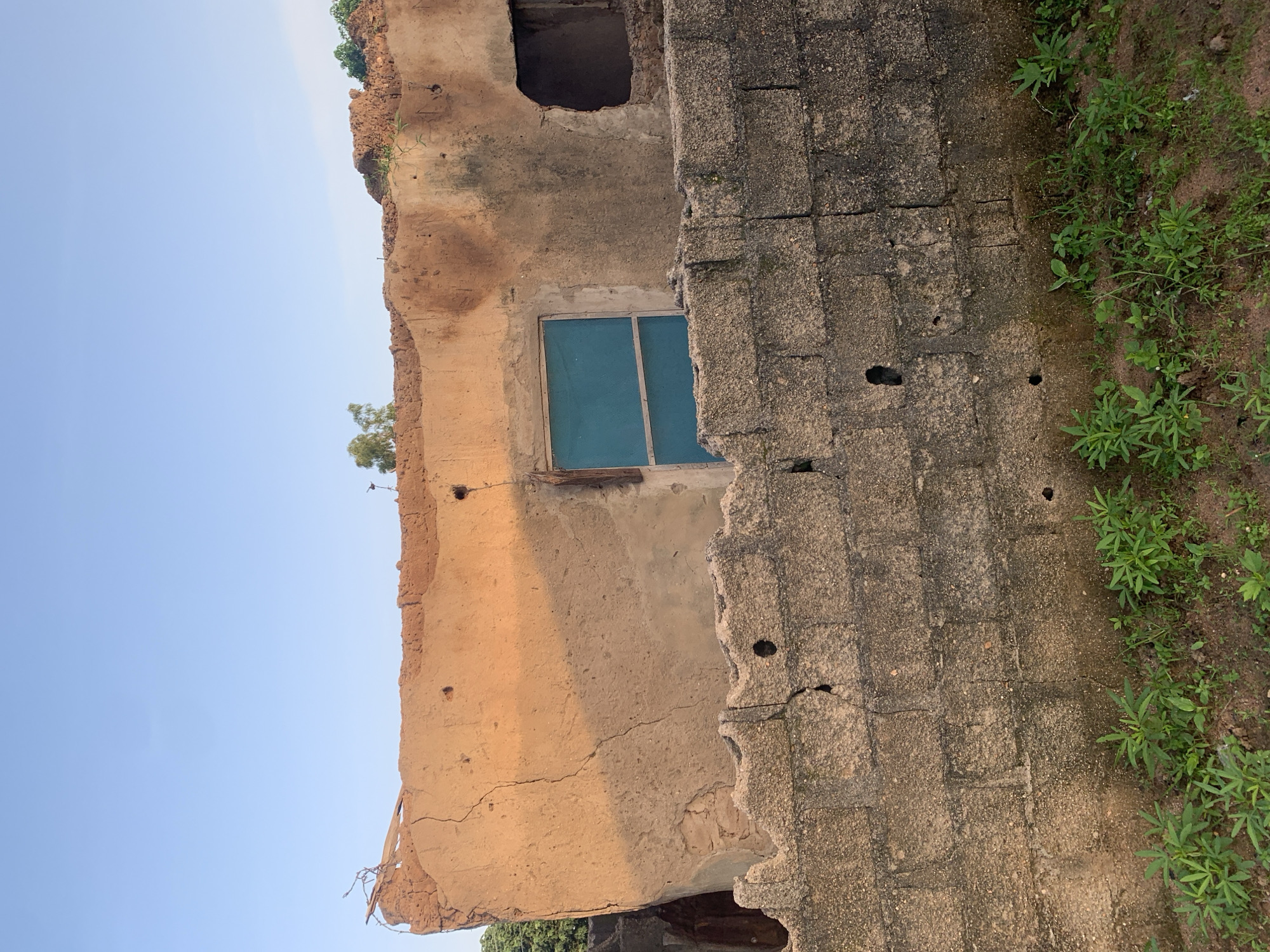 The image captures the exterior of a dilapidated building, possibly in the city of Jalingo, Nigeria. The structure appears to be constructed with mud or a similar earth-based material, showing significant signs of decay and erosion. The roofline is uneven and partially collapsed, with exposed earthen material and some scattered debris. A single window with a blue screen is visible on the facade, contrasting with the weathered texture of the wall. To the left of the window, there is an empty, dark opening that may have been another window or doorway. In the foreground, a rough stone wall, constructed from concrete blocks, runs horizontally across the image. This wall also exhibits signs of wear and tear, with cracks and missing sections. Behind the stone wall, a patch of green vegetation, consisting of small plants and grass, grows from the ground. The sky above is clear and a pale blue, indicating daytime. The overall impression is one of neglect and disrepair.