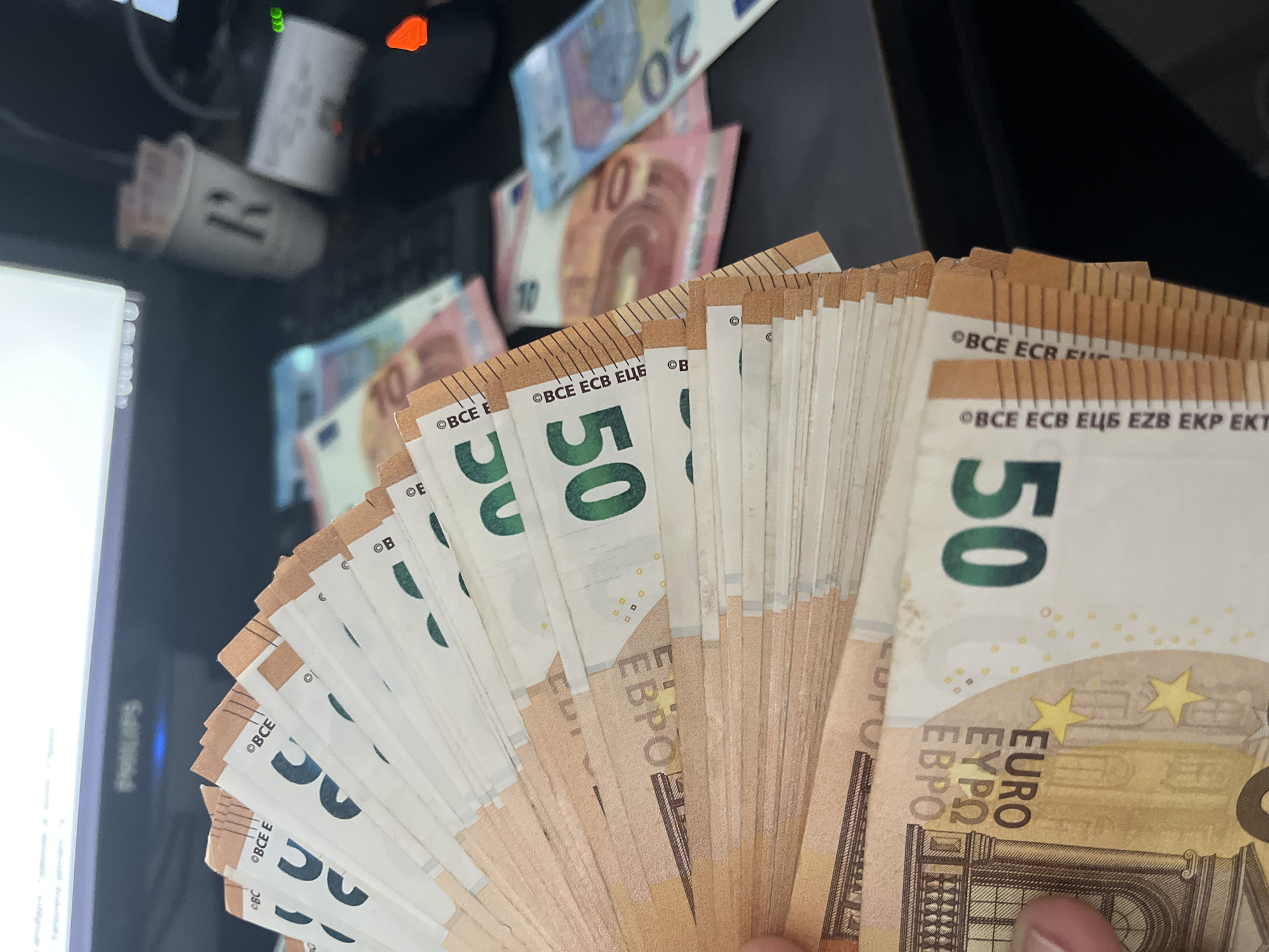 The image presents a close-up view of a person holding a large fan of 50 Euro banknotes, with more currency and electronic equipment blurred in the background.
In the foreground, a hand holds approximately 20 to 30 fifty Euro banknotes, fanned out to display their faces and edges. The prominent "50" denomination is visible in green, along with the currency names "EURO," "ΕΥΡΩ," and "EBPO." The edges of the banknotes clearly show security text variations such as "©BCE ECB EЦБ EZB EKP EKT," "©BCE ECB F," and "©BCE." The notes appear crisp and relatively new.
The background is dark and out of focus, featuring additional Euro banknotes of different denominations, specifically 10 Euro and 20 Euro notes, scattered on what appears to be a desk or table. Behind these notes, electronic devices are visible. There is a computer monitor, partially showing the brand name "Philips." Also visible is what seems to be a computer tower or another electronic device with green and orange indicator lights, and a white cylindrical object with a black marking that resembles "R" or "PR." The overall setting is indoors, likely a home or office environment. The scene suggests the display of a considerable amount of cash. The location context indicates Ivailo, Bulgaria.