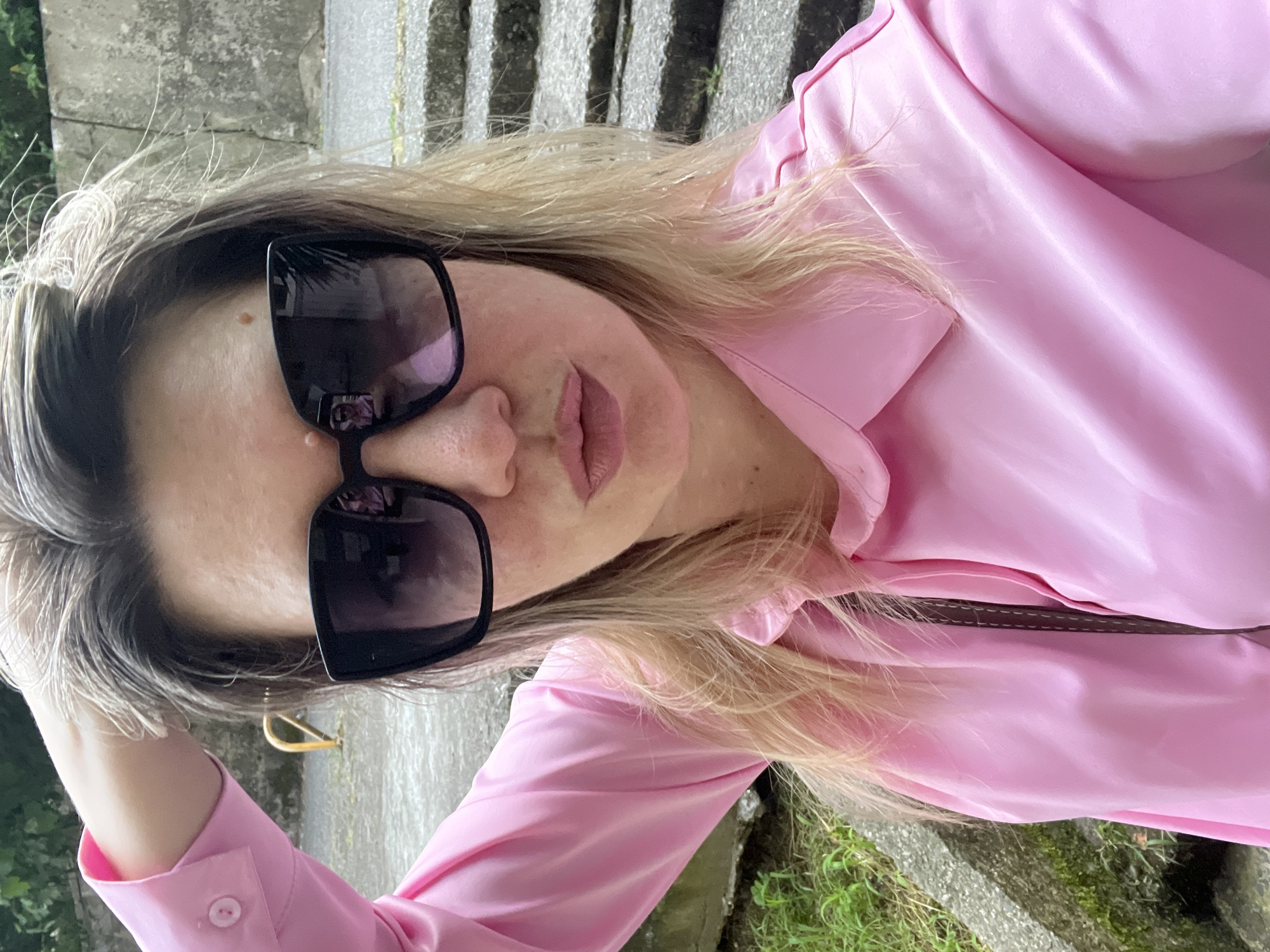 In an outdoor urban setting, a woman with blonde hair and sunglasses poses for a selfie. She is wearing a pink collared shirt and her right hand is raised, touching her hair. The background shows concrete steps, greenery, and what appears to be a yellow metal railing. The image was taken in Sochi, Russia.