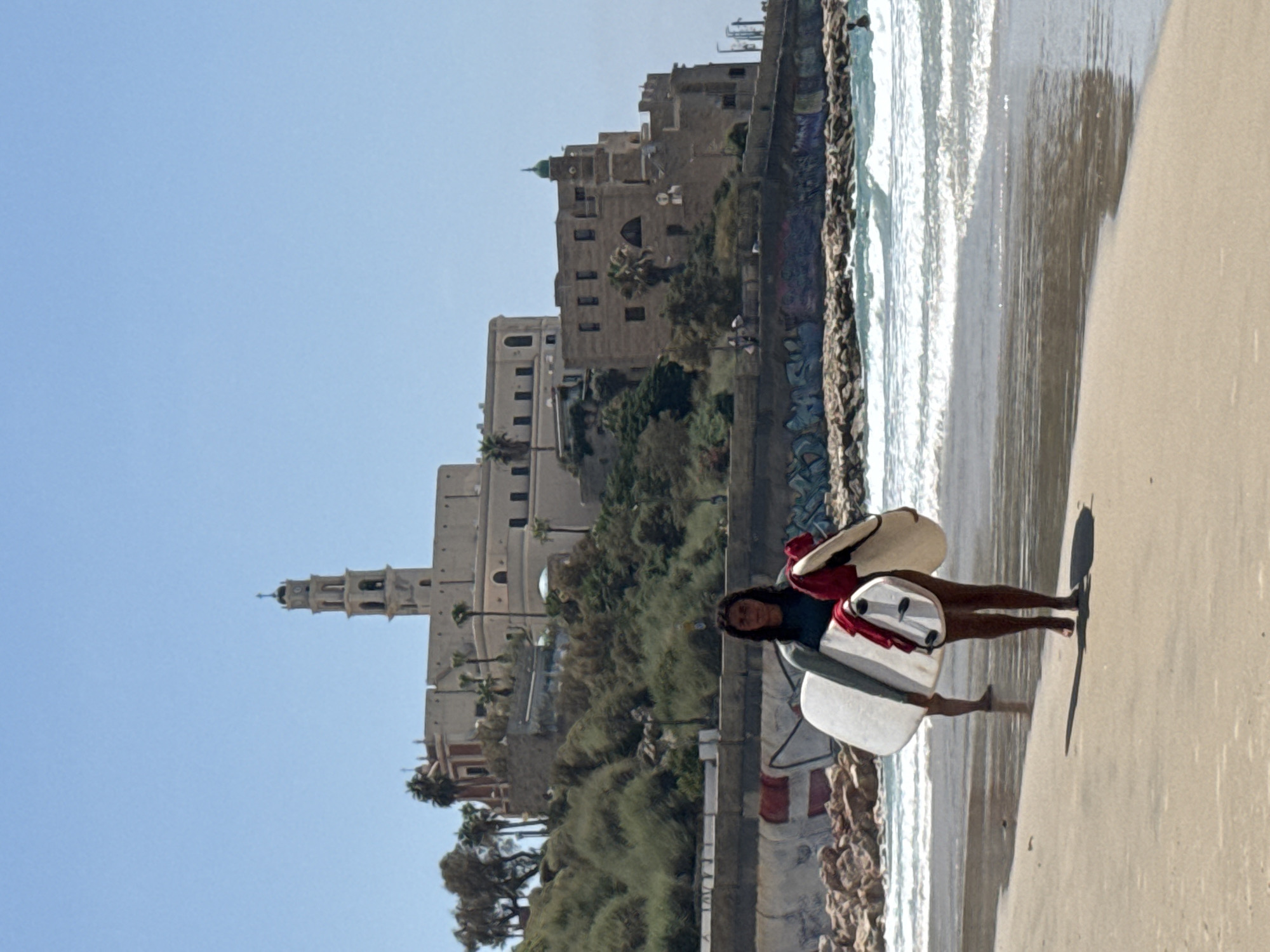 The image depicts a sunny day at the beach in Tel Aviv, Israel, with the historic port of Jaffa visible in the background. A young woman, wearing a wetsuit, walks along the sandy shore carrying two surfboards. She is facing the camera and her expression is neutral. Behind her, another person is partially visible, also on the beach. The sea is calm with gentle waves, and a few people can be seen in the water. The architecture of Jaffa, with its characteristic stone buildings and a tall bell tower, stands out against the clear blue sky. The coastline is lined with a breakwater made of rocks, some of which are covered in graffiti. Palm trees and other vegetation dot the landscape leading up to the buildings. The overall scene conveys a sense of leisure and outdoor activity on a beautiful day.