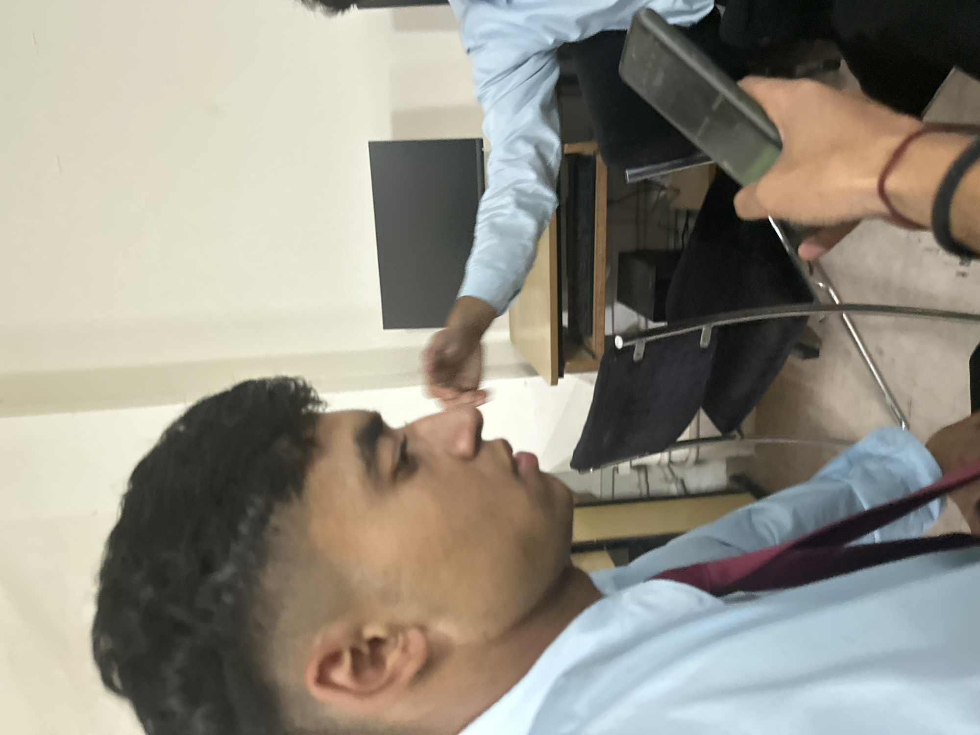 The image shows a close-up, side-profile view of a young man with dark, short, curly hair styled with an undercut fade. He is wearing a light blue collared shirt and a dark red tie. His expression is neutral as he looks downwards. In the background, another person's arm, wearing a light blue shirt, is visible, holding a dark-colored smartphone. The environment appears to be an indoor setting, possibly an office or classroom, with a computer monitor on a desk and chairs. The lighting is neutral, suggesting an interior space with artificial light. The location is Unknown, Unknown.
