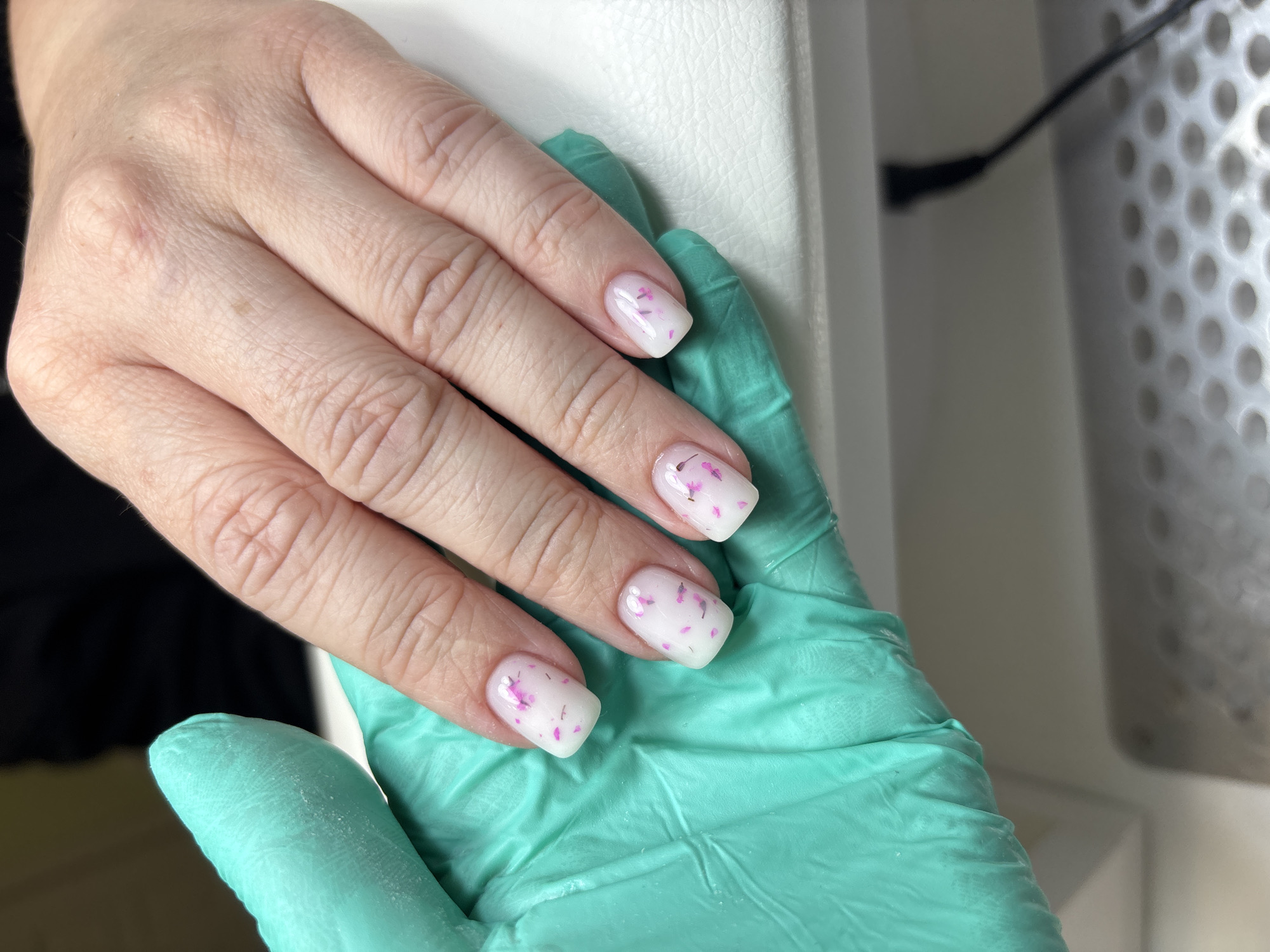 A close-up shows a person's hand wearing disposable turquoise gloves, holding another hand. The hand being held has manicured nails with a white base and small pink floral designs. The background consists of a white surface and part of a black cable connected to a device with a perforated white cylindrical shape. The location is indoors.