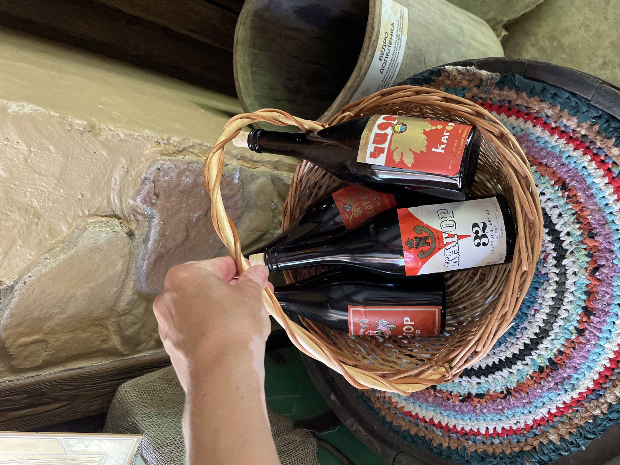 A hand is holding a woven basket filled with four dark glass bottles, likely wine or spirits. The basket is made of natural wicker and has a braided handle. Each bottle has a red and yellow label. One label, on a bottle at the top of the basket, features a golden image of grapes and reads "КАГОР". Another label, partially visible, also displays red and gold elements and appears to be for a similar product. The basket sits on top of a dark wooden barrel, which is covered with a colorful, handmade rag rug featuring stripes of red, blue, white, purple, and black. In the background, a large, dark, vintage-looking wooden vessel or barrel is visible. To the left, a textured, light-colored wall with visible brickwork peeks out. The overall scene suggests a rustic setting, possibly a winery or a traditional establishment in Estosadok, Russia. The lighting appears to be natural, hinting at daytime. There is text on one of the labels that reads "КАГОР", and a partial label in the background shows Cyrillic text, likely including "ВЕДРО ДОБЛЕПЕНКА".