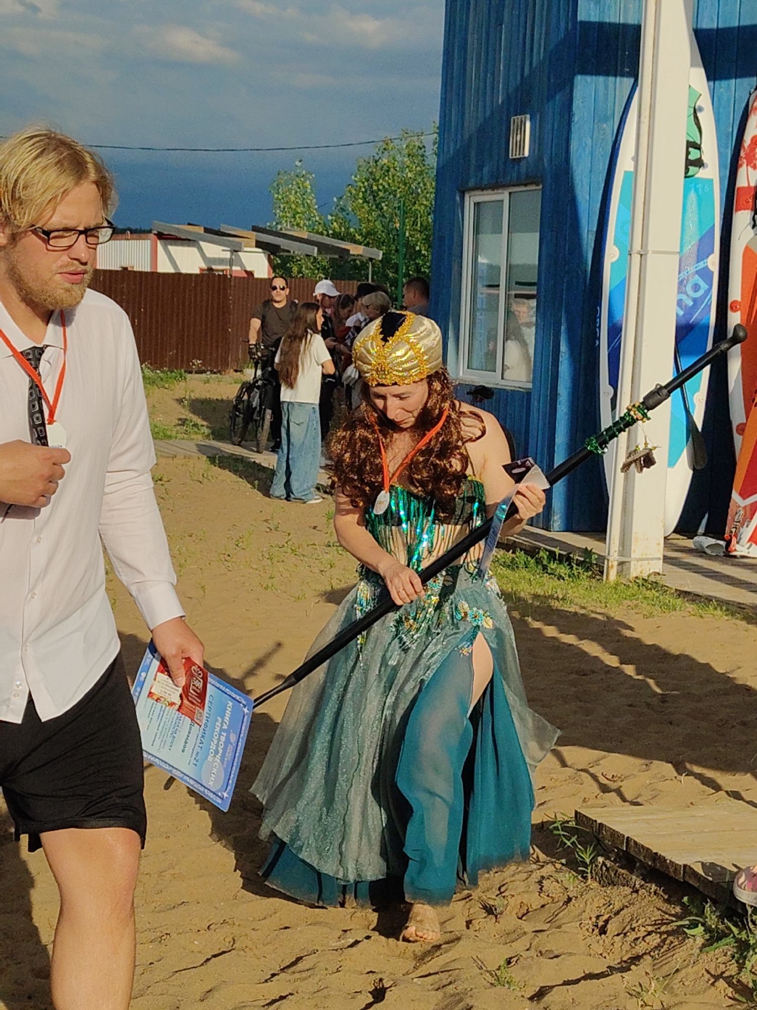 An outdoor medium shot captures a woman in a turquoise, sequined costume adorned with a golden headdress, holding a black staff with green decorations and charms. She is standing on a sandy area, with a blue wooden building and several surfboards in the background. To her left, a man wearing glasses, a white shirt, and black shorts is visible, holding a blue certificate. Other people and a bicycle are also present in the background. The scene appears to be set in a recreational area with a beach-like environment.