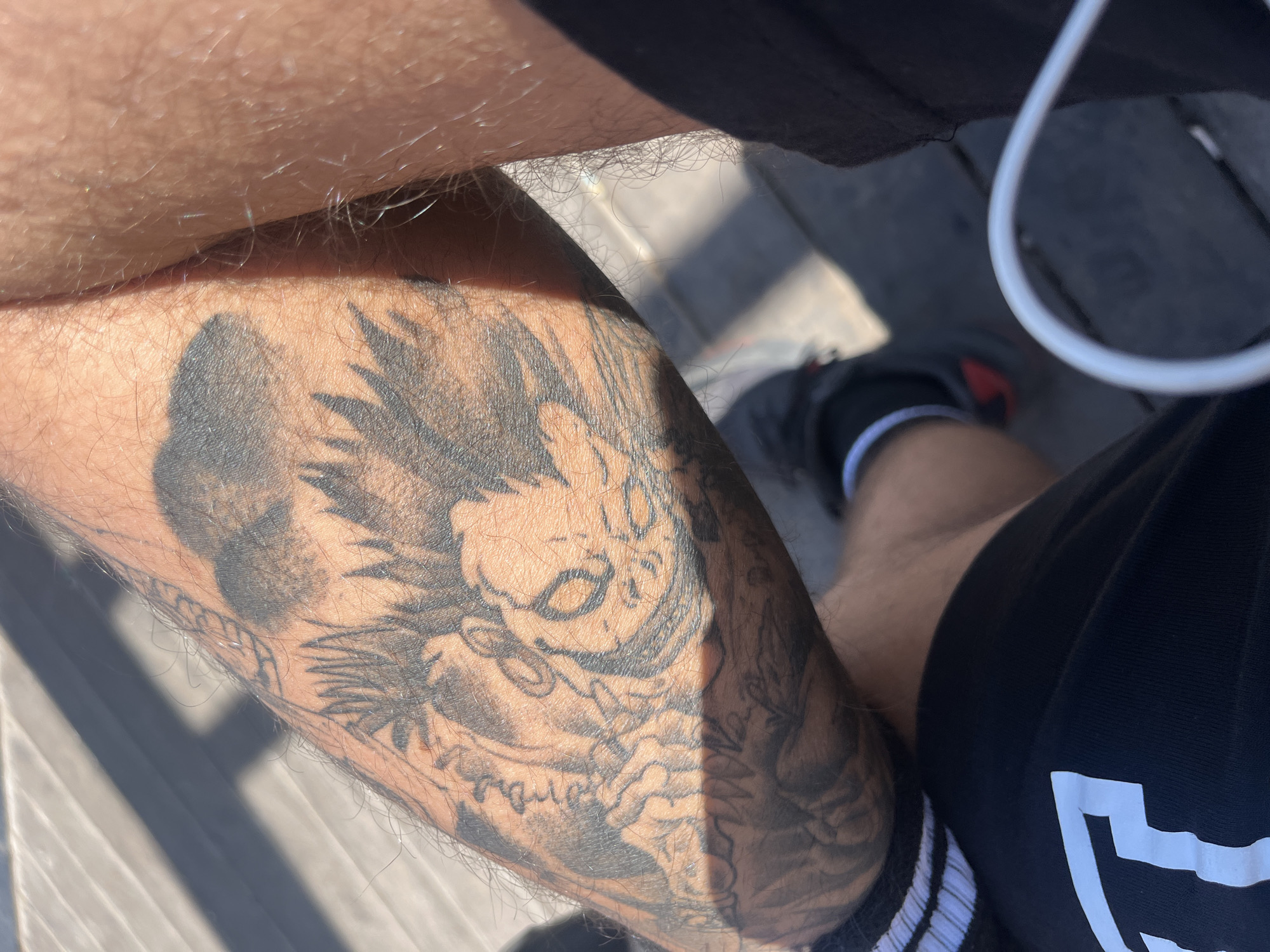 A close-up, high-angle shot features a person's left forearm and part of their thigh. A large, detailed black and grey tattoo covers a significant portion of the forearm. The tattoo appears to depict a stylized character, possibly from animation or fantasy. The person is wearing black shorts with white stripes near the hem, and a white earbud cord is visible. A sneaker-clad foot is also partially visible. The setting seems to be outdoors, possibly on a partially-shaded wooden structure. The location is indiscernible, and nothing in the image indicates that it is in Arica, Chile.