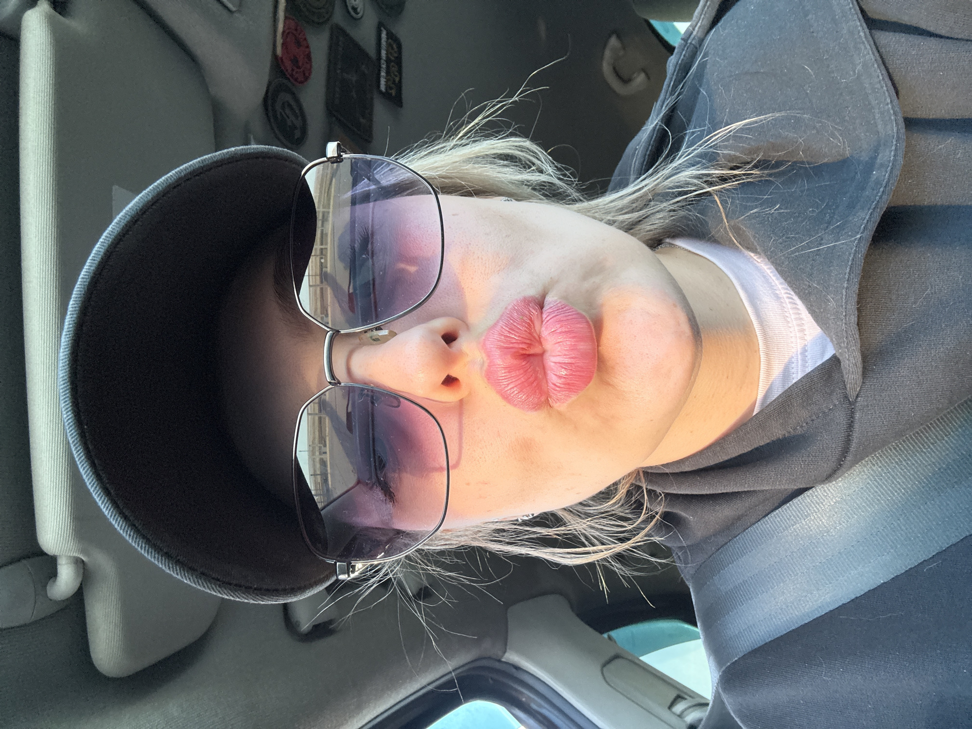 A woman is taking a selfie inside a car. She is wearing sunglasses and a visor. She is also wearing a dark-colored hooded sweatshirt and a seatbelt. The car's interior is gray. Patches are affixed to the car's visor. The background suggests an outdoor environment with ambient lighting. It is not possible to identify the location as Sherlovaya Gora, Russia from this image.