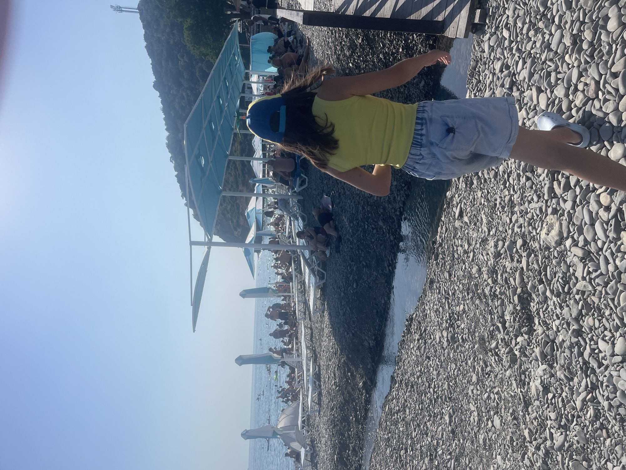 The image depicts a crowded beach scene on a sunny day. In the foreground, a young person wearing a yellow sleeveless top and denim shorts walks away from the camera, heading towards a wooden walkway on the right. They are wearing a blue and yellow baseball cap. The ground is covered in smooth, grey pebbles. To the left, a narrow stream of water flows towards the sea.
The beach itself is lined with numerous white lounge chairs and blue and white umbrellas, suggesting a resort or public beach. Many people are visible in the background, seated on the chairs, walking, and swimming in the calm, blue ocean. The water is clear enough to see some individuals wading or floating.
In the distance, a lush, green hillside rises, topped with trees and a cell tower. The sky is a clear, bright blue, indicating a cloudless day. The overall atmosphere is one of a busy, recreational beach environment. The location is identified as a beach in an unspecified city.