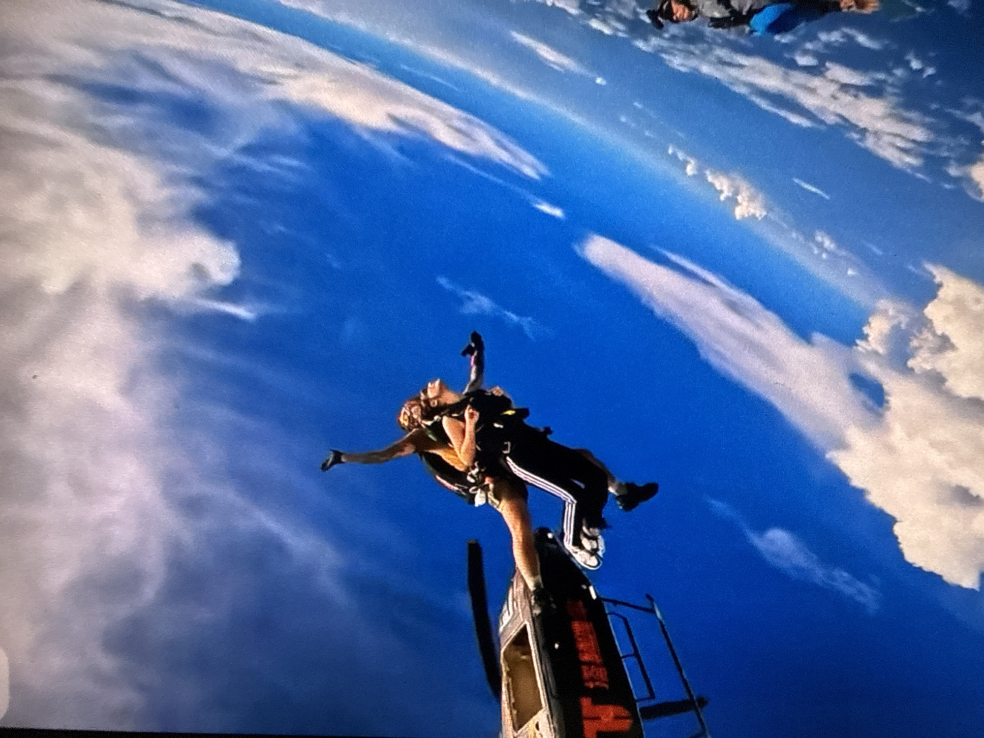 A low-angle shot shows two skydivers exiting a helicopter against a bright blue sky with scattered white clouds. One person is visible at the upper right, also appearing to be skydiving. The helicopter, only partially visible, is orange and dark gray with a ladder on its side. The two skydivers are connected with gear. The person closer to the helicopter is wearing shorts and the person above is wearing dark pants with white stripes.