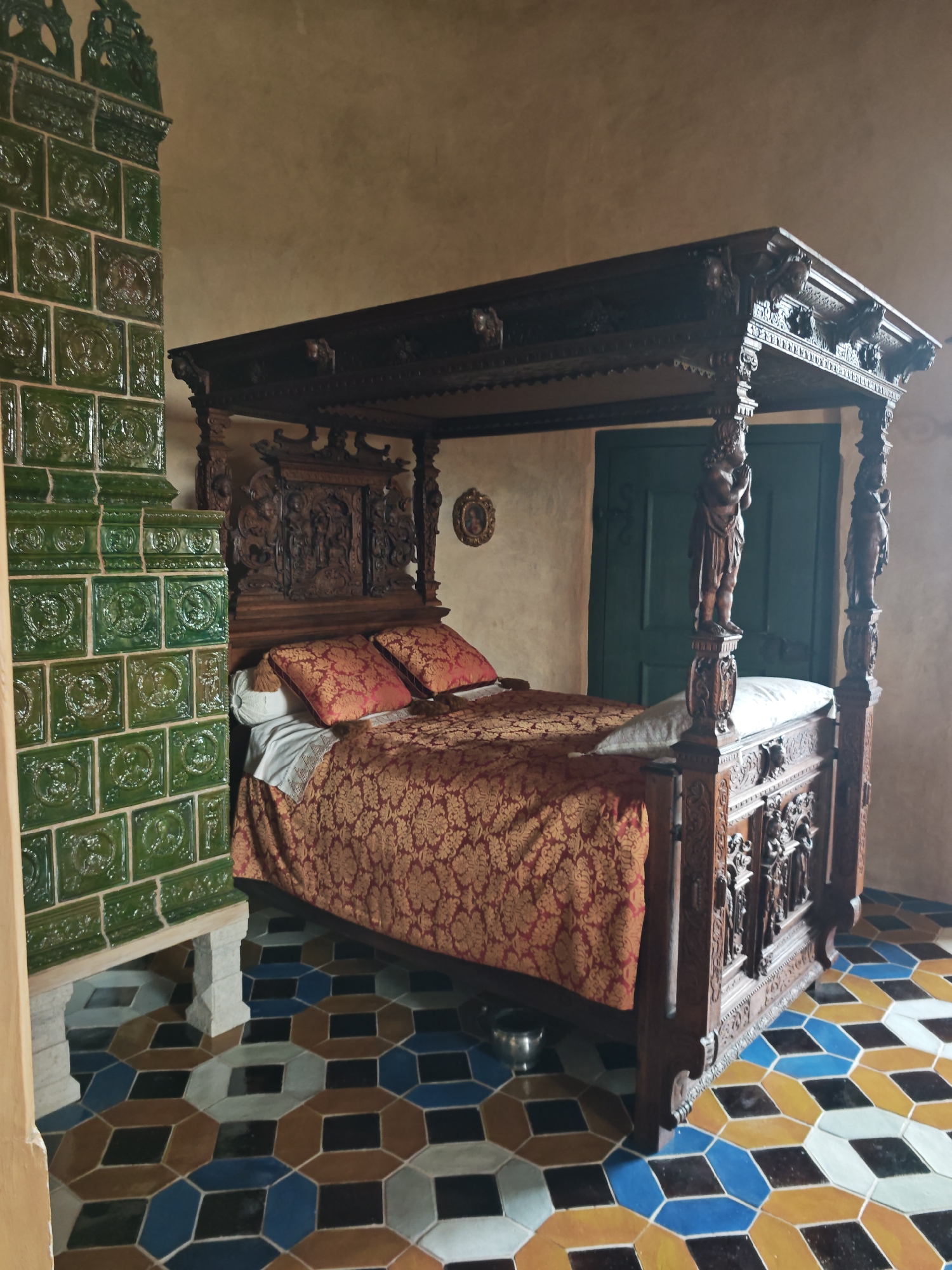 This is a medium-shot interior photograph of a bedroom in Bauska Castle, Latvia, likely a historical exhibit room. The focal point is a large, ornate four-poster wooden bed with intricate carvings on the posts and headboard, draped in a richly patterned red and gold fabric. Two red patterned pillows rest at the head of the bed. To the left of the bed is a tall, green tiled stove with decorative relief patterns. The floor is covered in a colorful geometric tile pattern of blues, browns, grays, and yellows. In the background, a closed dark green door is visible. The walls are a muted, textured tan color.