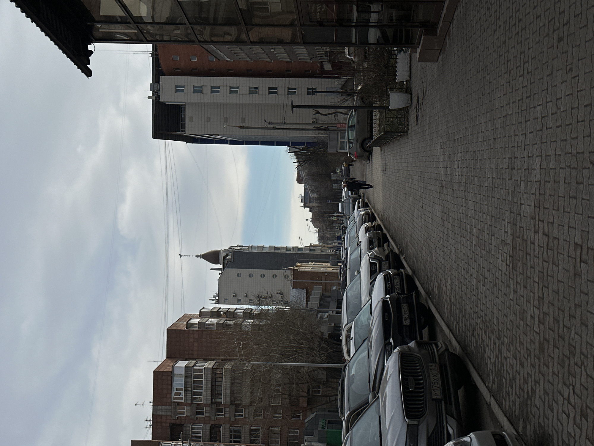 A long row of parked cars lines a brick-paved street in Perm, Russia, near several multi-story buildings. The architecture of the buildings ranges from older brick apartments to modern, white-faced buildings. The sky is overcast with thin clouds, suggesting the photo was taken during the daytime. A few people are walking further down the street away from the camera. Streetlights and bare trees line the sidewalk. License plates on the vehicles indicate they are registered in Russia.
