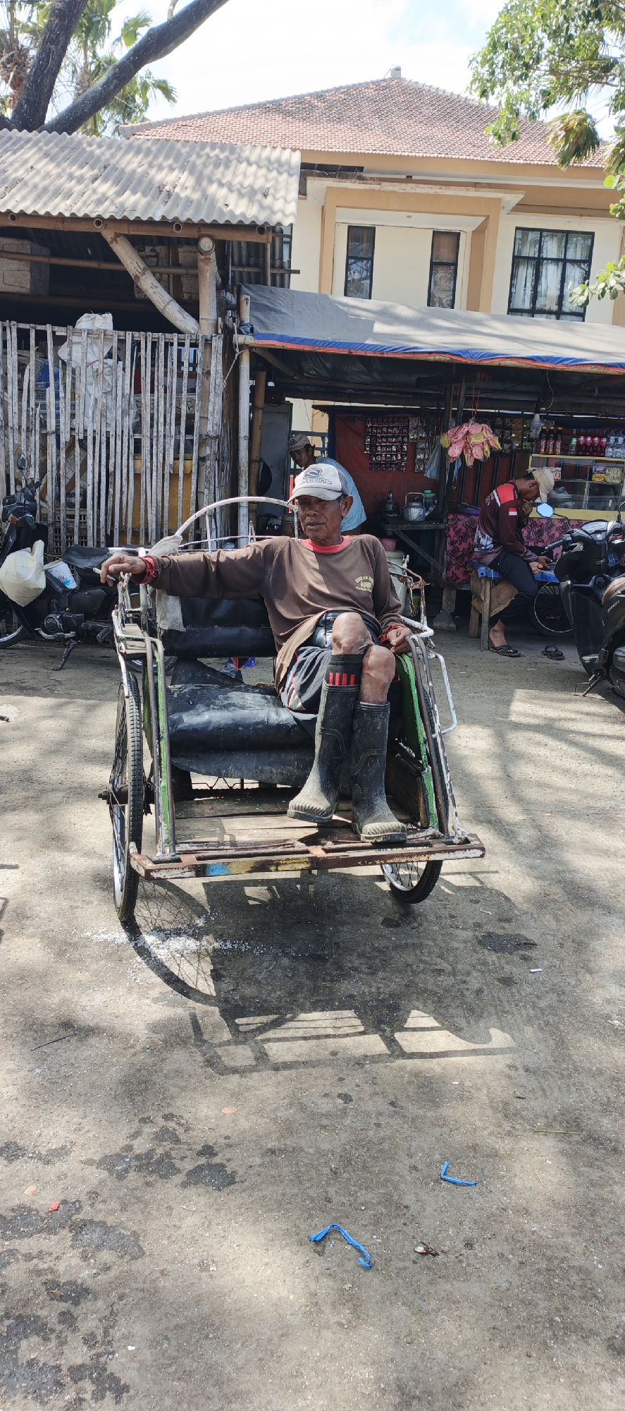 An elderly man wearing a brown long-sleeved shirt, dark shorts with red stripes, and tall black rubber boots sits in a pedicab. He wears a white cap. The pedicab has a green and black frame and appears worn. In the background, a building with a red tile roof stands behind a row of small shops. A motorbike is parked to the left, and another motorbike is parked to the right of the shops. The ground is paved with concrete and appears dirty. A blue plastic tie lies on the ground in the foreground. The scene is set in the city of Puger, Indonesia.