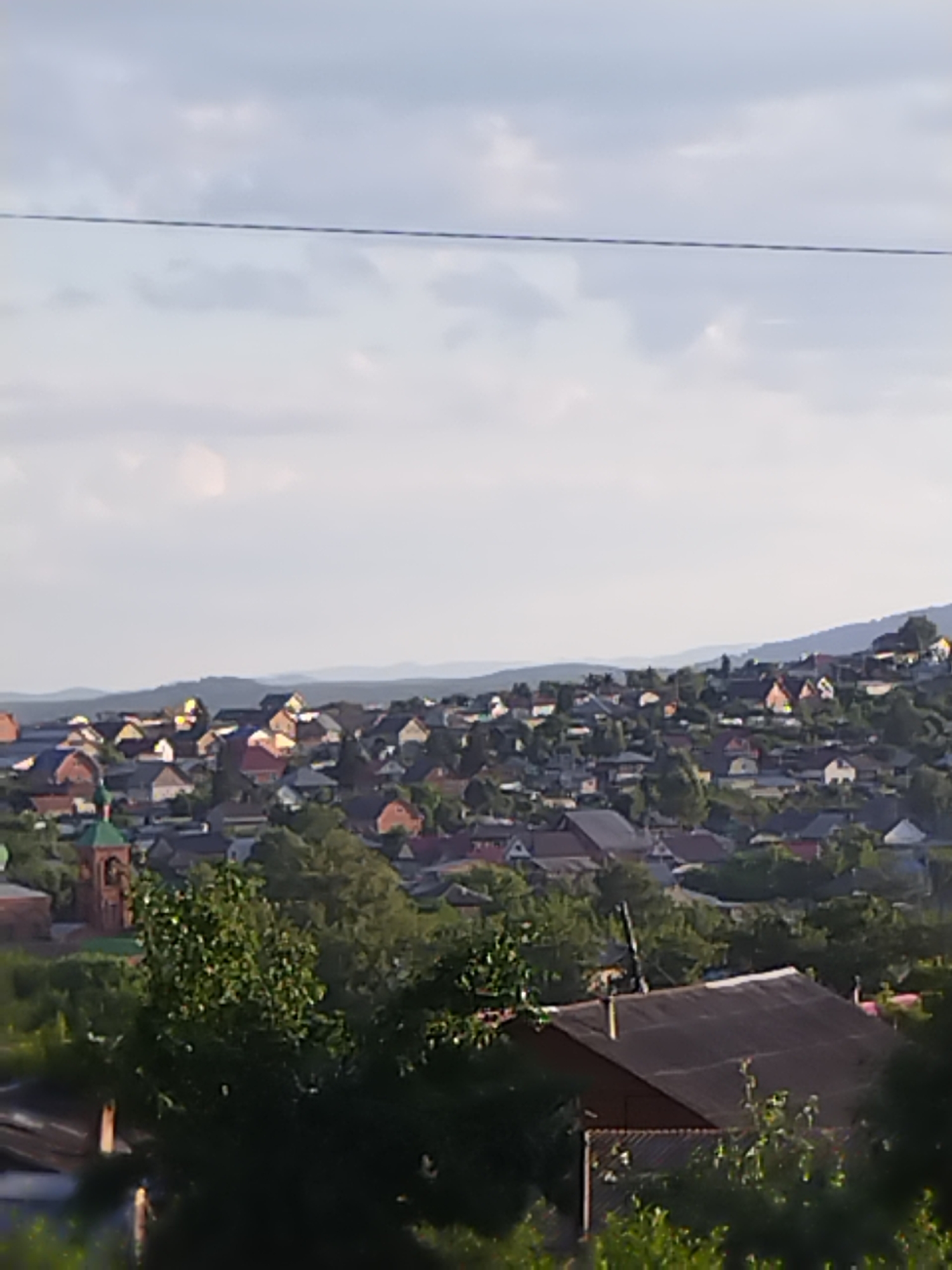 The image depicts a panoramic view of a residential area nestled on a hillside under a cloudy sky. The foreground is dominated by lush green trees, with a large, dark-roofed building partially visible in the lower right corner, obscured by foliage. Beyond the trees, a dense collection of houses with varied roof colors, including red, brown, and gray, spreads across the incline. A prominent brick building with a green dome and spire stands out on the left side of the town. The background features rolling hills and a hazy horizon, suggesting a rural or semi-rural setting. The overall impression is one of a quiet, established community situated within a natural landscape.