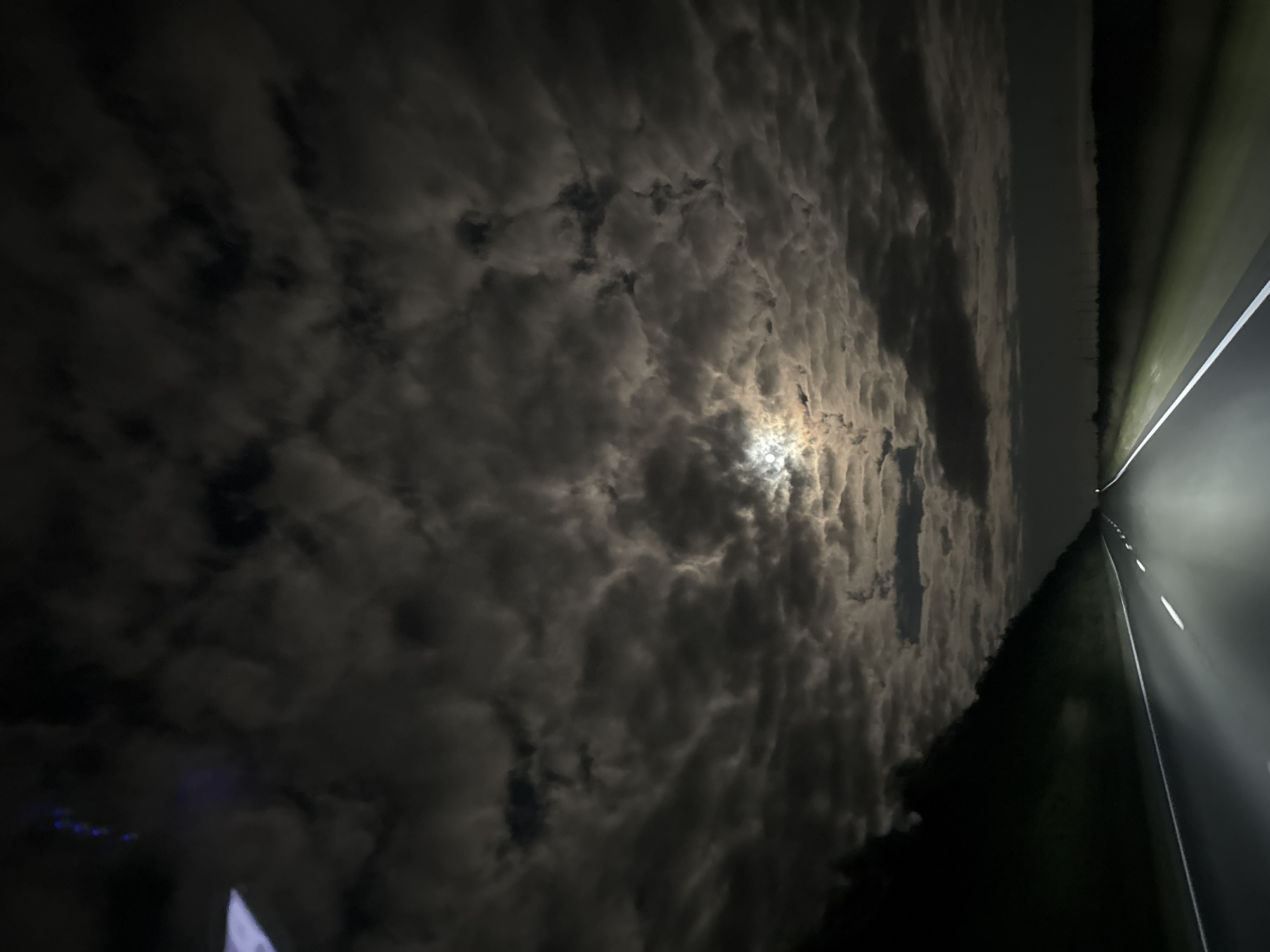 The image depicts a nighttime view of a highway, likely in or near Михайловка, Russia. The road is paved with white lane markings illuminated by headlights. To the left of the road, there is a dark, tree-lined embankment. The sky is filled with dramatic, thick clouds, illuminated by a bright moon peeking through a gap. The overall atmosphere is dark and somewhat ominous, with the headlights cutting through the gloom. There are no visible people or vehicles other than what is implied by the presence of the highway and headlights. The horizon in the distance is obscured by darkness and possibly more trees or distant lights, though it's difficult to discern specific details.