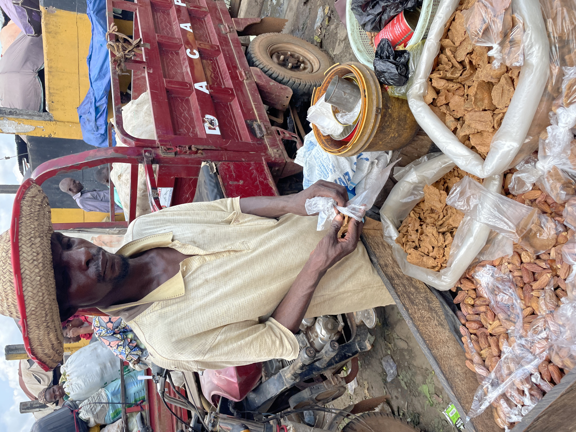 A medium shot shows a man wearing a straw hat and a light-colored, collared shirt with the collar popped. He is standing in front of a wooden crate filled with what appear to be dried dates and dried, processed meat or snacks. His right hand is holding a clear plastic bag and a piece of the processed food item. In the background, a red flatbed truck is visible, with "A C A P" written on its side. Other people, bags of goods, and market stalls are also present in the background, suggesting a market environment. The ground is dirt and debris.