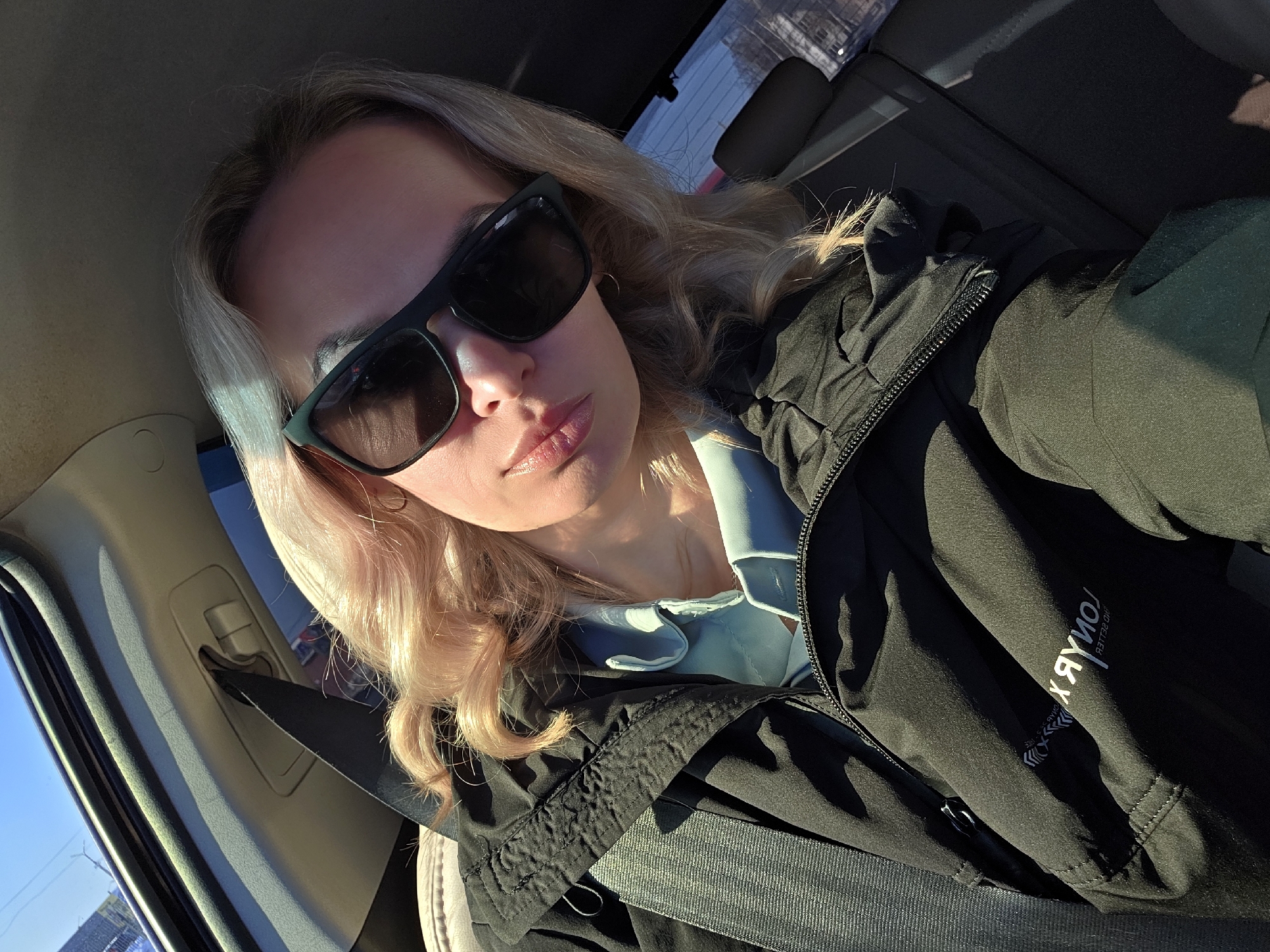 A female individual with blonde hair, wearing dark sunglasses, a light blue collared shirt, and a dark zippered jacket, is depicted in a selfie taken inside a vehicle in Chaikovsky, Russia. A dark seatbelt strap crosses her chest, originating from the vehicle's upper-left pillar, which has a light-colored headliner. Sunlight illuminates the subject's face from the upper right. The jacket features a partially mirrored text logo on its left sleeve. Through the vehicle's windows, a clear blue sky is visible, along with blurred distant urban or suburban elements, including what appears to be a utility pole or antenna in the upper left background.