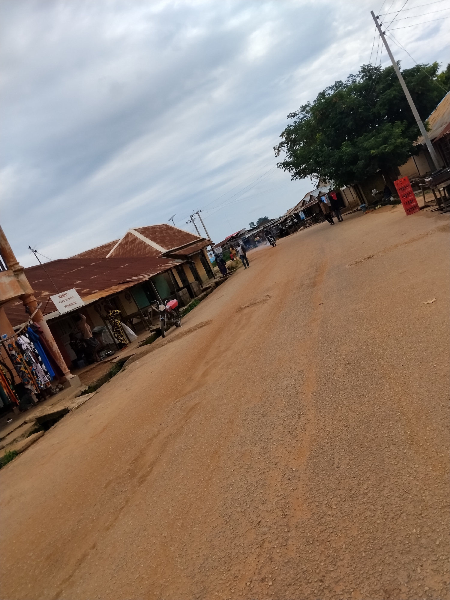 In Mangu Town, Nigeria, a dusty, unpaved road runs through a market area lined with shops featuring corrugated metal roofs. On the left, a storefront displays colorful clothing on hangers. A red and white sign above the storefront reads "NWANK'S CAKE N' MORE 08140384157". A motorcycle is parked in front of one of the shops. Several people are visible walking along the road and in front of the shops. A large tree with dense green foliage stands on the right side of the road, next to a utility pole and wires. In the background, more buildings and trees suggest a semi-urban environment. The sky is overcast with clouds.
