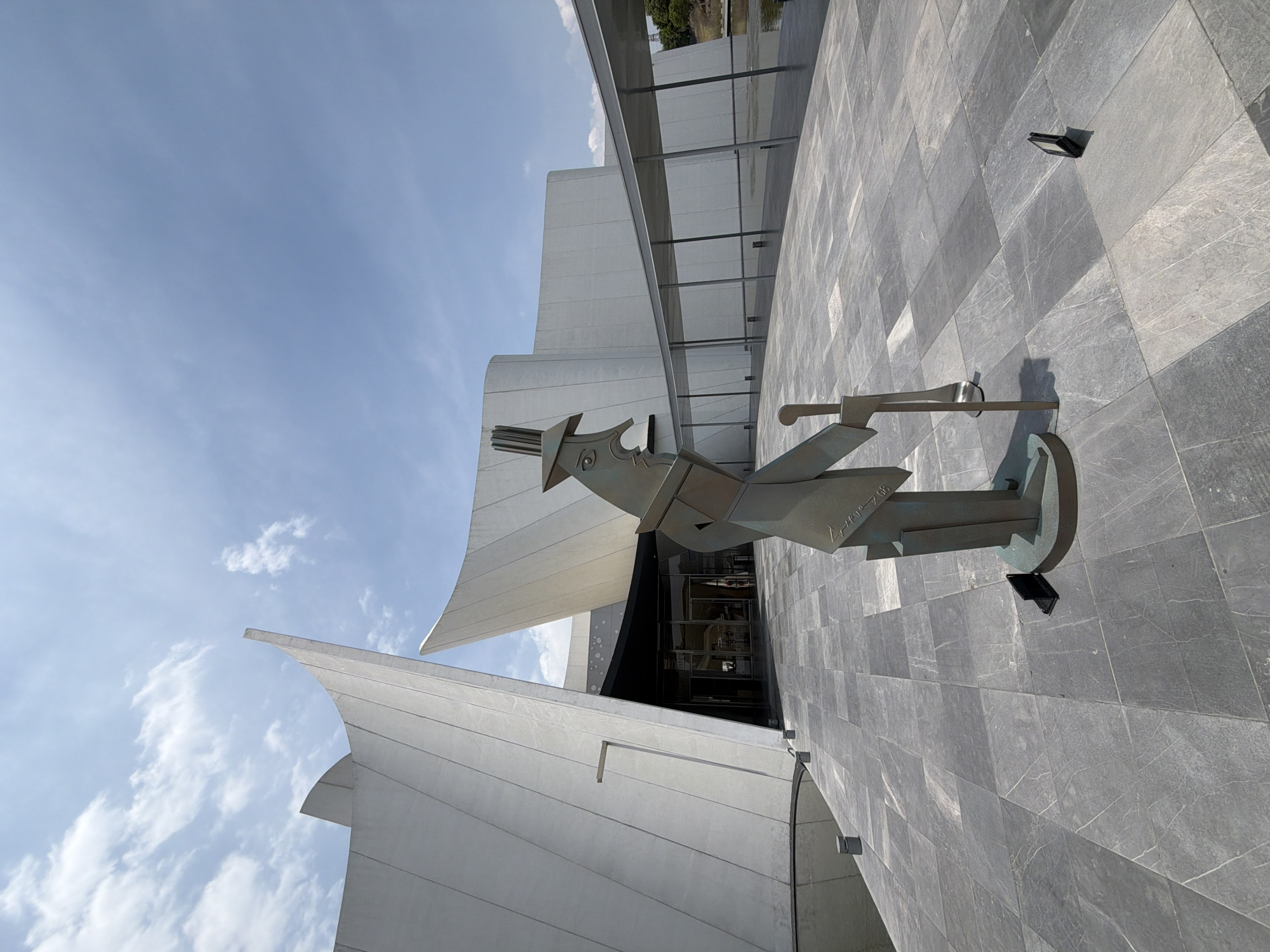 A low-angle shot showcases a sculpture of a person in front of a modern architectural building against a bright blue sky. The sculpture is made of a metallic material, resembling bronze or a similar alloy. The figure is stylized with geometric shapes and appears to be holding a long object or instrument. The building behind the sculpture has white, sail-like structures and a gray tiled exterior, suggesting a museum or cultural center. A close inspection of the sculpture reveals illegible writing on the side. The shot appears to have been taken in San Bernardino Tlaxcalancingo, Mexico.