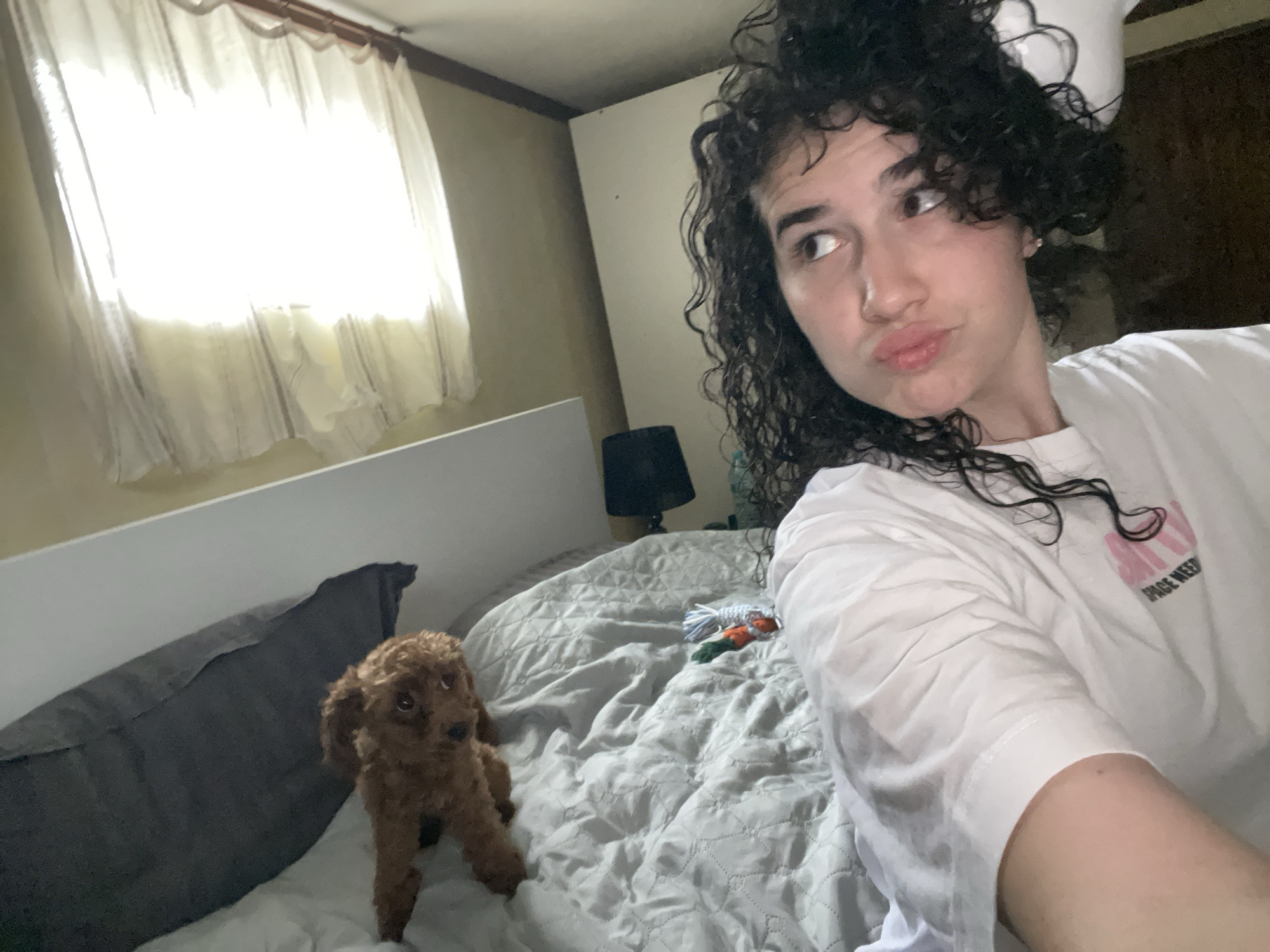 A woman with curly hair is taking a selfie in what appears to be a bedroom. She is wearing a white shirt and looking off to the side with a pouting expression. A small, brown poodle is standing on the bed next to a gray pillow and gray bedspread. A window with a white curtain is visible in the background, along with a black lamp. The location is not discernible in this image, therefore it is impossible to determine if this photo was taken in Sofia, Bulgaria.