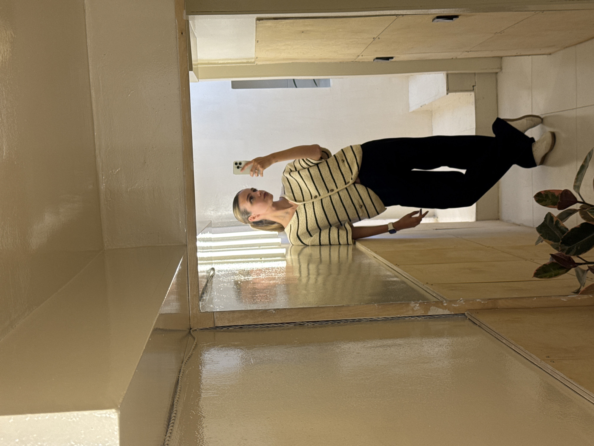 The image shows a young woman taking a selfie in an indoor setting, likely a corridor or spacious room. She has blonde hair pulled back and is wearing a cream-colored, horizontally striped cardigan over a lighter top, black wide-leg pants, and light-colored shoes. She holds a light-colored smartphone in her right hand, pointed towards herself, with a focused, neutral expression. A watch is visible on her left wrist.
The setting features light-colored walls and a distinctive floor. The floor is composed of large, light-toned panels, some of which are highly glossy and reflective, appearing wet or freshly coated with a self-leveling resin or similar material. These reflective sections show clear reflections of the ceiling and parts of the woman's body. Adjacent to these are matte, lighter-colored panels, possibly unfinished wood or a different flooring type, creating a textural contrast. Thin dark lines mark the seams or expansion joints between the floor panels.
On the far right, a plant with large, dark green and reddish leaves, resembling a Ficus Elastica, is visible, adding a natural element to the otherwise clean and possibly minimalist or under-renovation space. The lighting is bright and even, indicating daytime. No visible text is present in the image. The scene suggests a modern or recently updated interior, possibly in Tbilisi, Georgia, as per the context.